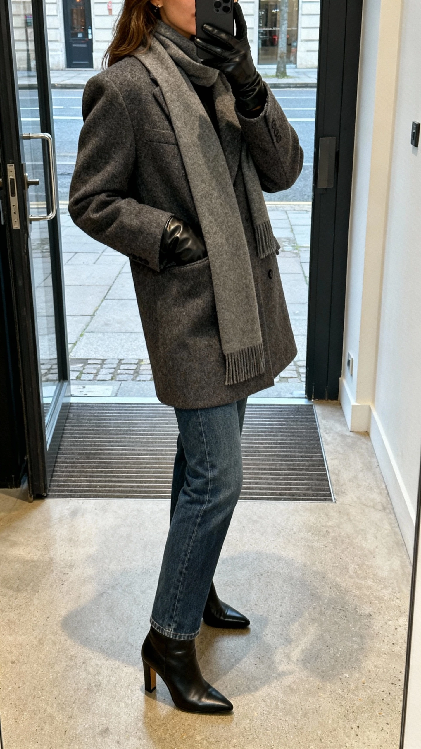 iPhone side-angle selfie showing a woman in a structured wool coat with a slim scarf neatly draped, straight-leg jeans, leather gloves, heeled boots, face not visible, city entryway, natural daylight, casual iPhone photo.