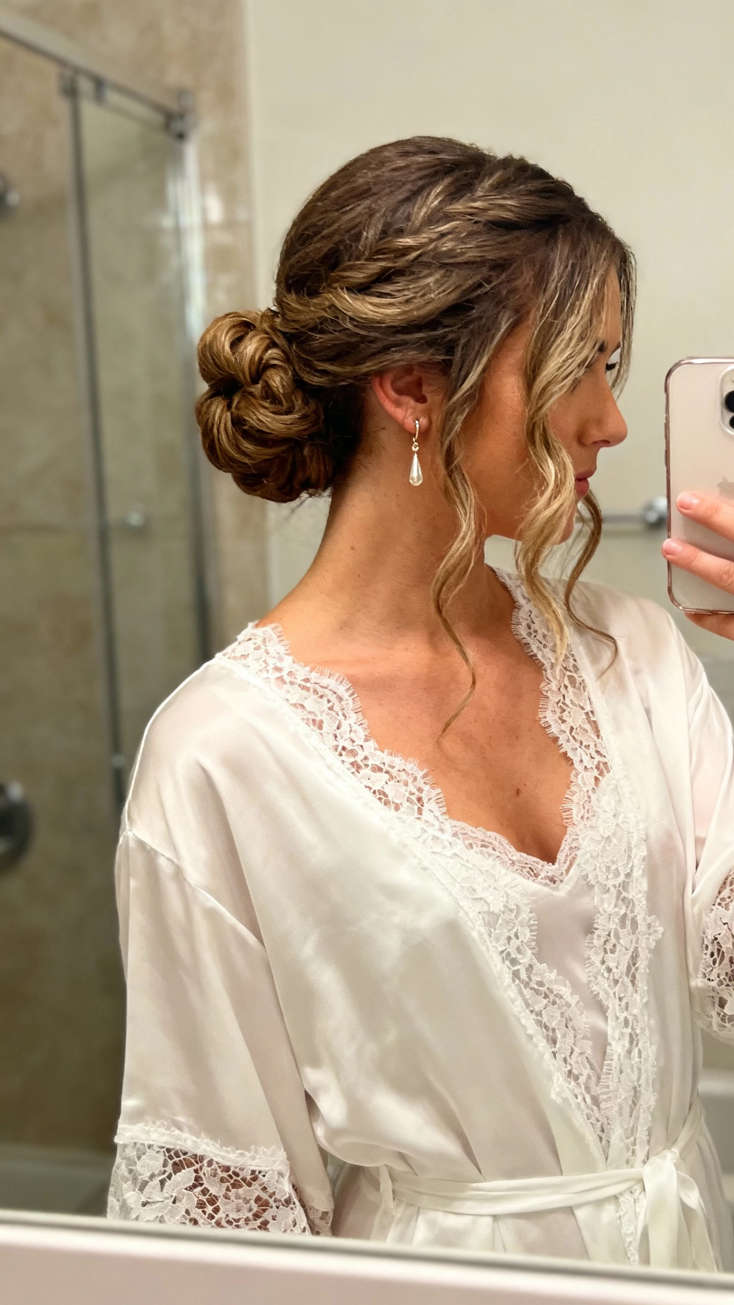 iPhone side-angle selfie showing a woman with a romantic textured low bun and soft face‑framing waves, dainty drop earrings, lace-trim bridal robe; face not visible, shoulder crop, bathroom mirror, warm indoor light, natural iPhone aesthetic.