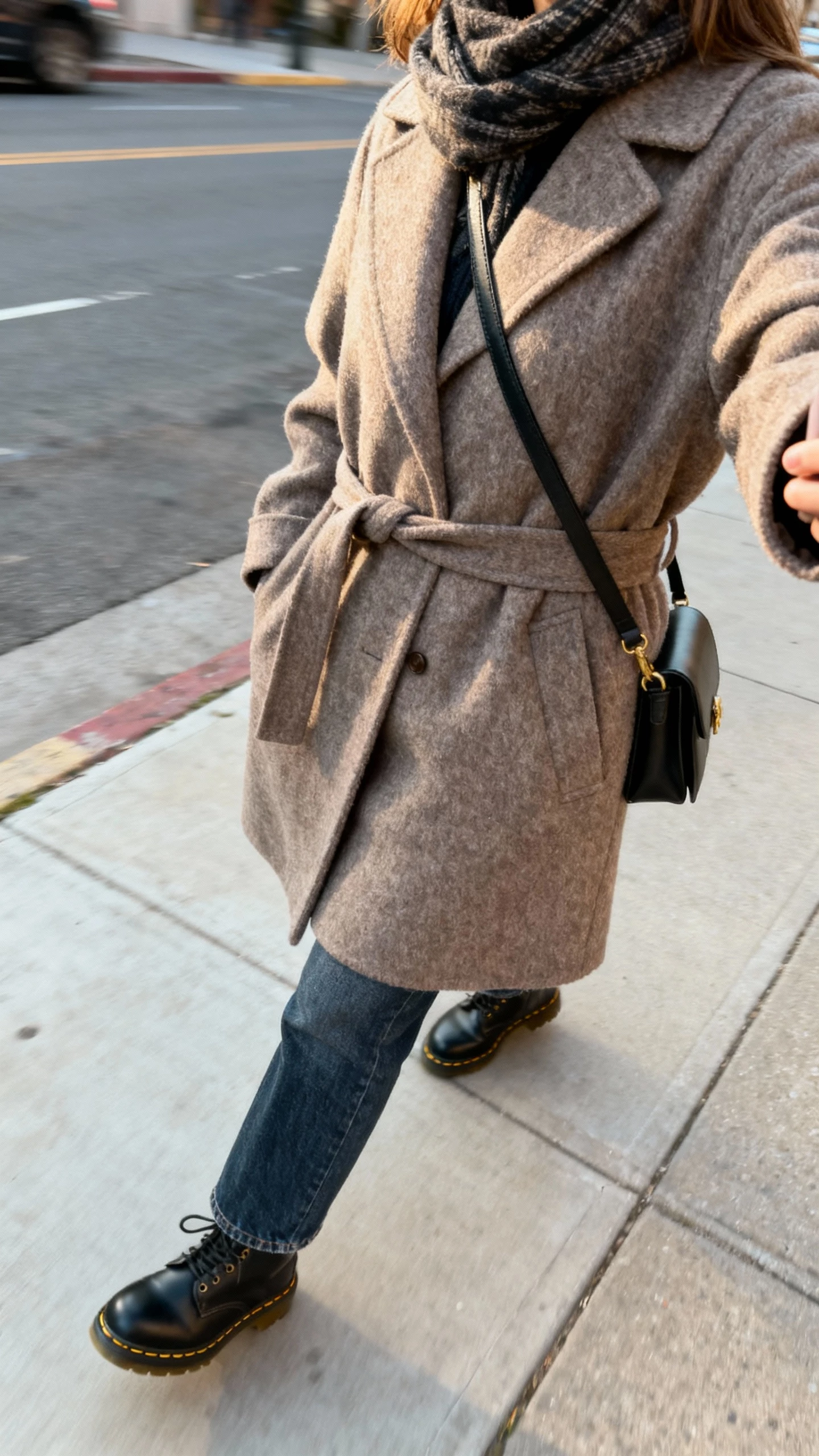 iPhone side-angle street selfie of a woman wearing a belted wool coat over straight-leg denim with Doc Martens, crossbody bag, scarf, face not visible, urban sidewalk in natural daylight, casual iPhone photo.