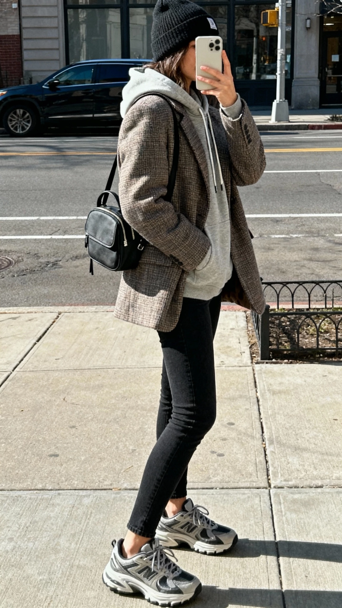 iPhone side-angle street selfie showing a woman wearing a winterized blazer layered over a hoodie, slim black jeans, chunky sneakers, beanie, mini backpack, face not visible, city sidewalk in crisp daylight, natural iPhone photo quality.