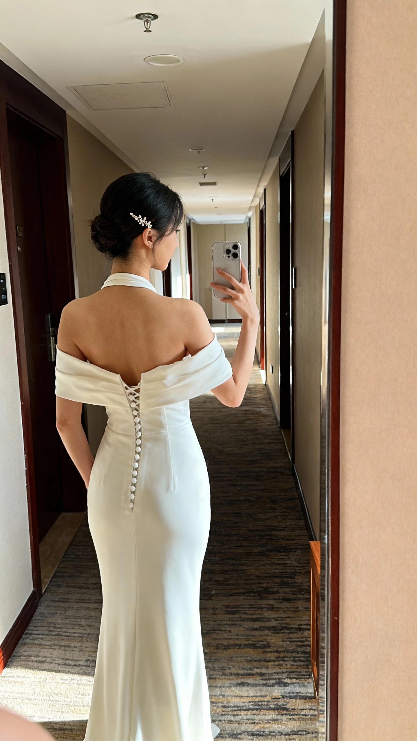 iPhone side-back selfie of a woman wearing a halter/high-shoulder wedding dress that lifts and highlights shoulders, open back with button detail, sleek column silhouette, dainty hairpin, face not visible, hotel hallway mirror, warm indoor lighting, natural iPhone photo.