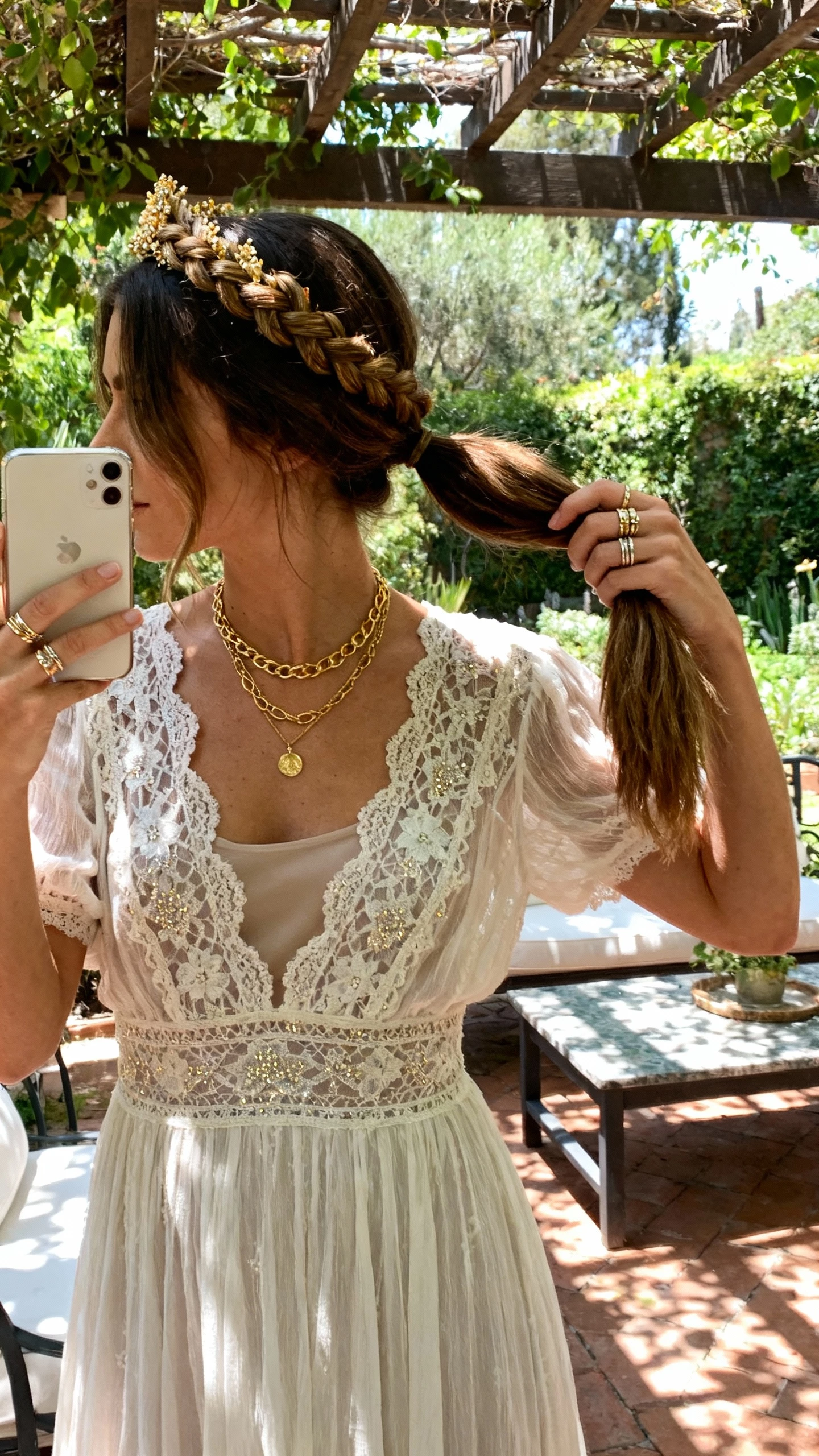 iPhone side/back selfie of a woman with a braided crown feeding into a ponytail, lace boho bridal dress, layered gold necklaces, stacked rings, face not visible, garden patio setting with dappled sunlight, natural iPhone photo quality