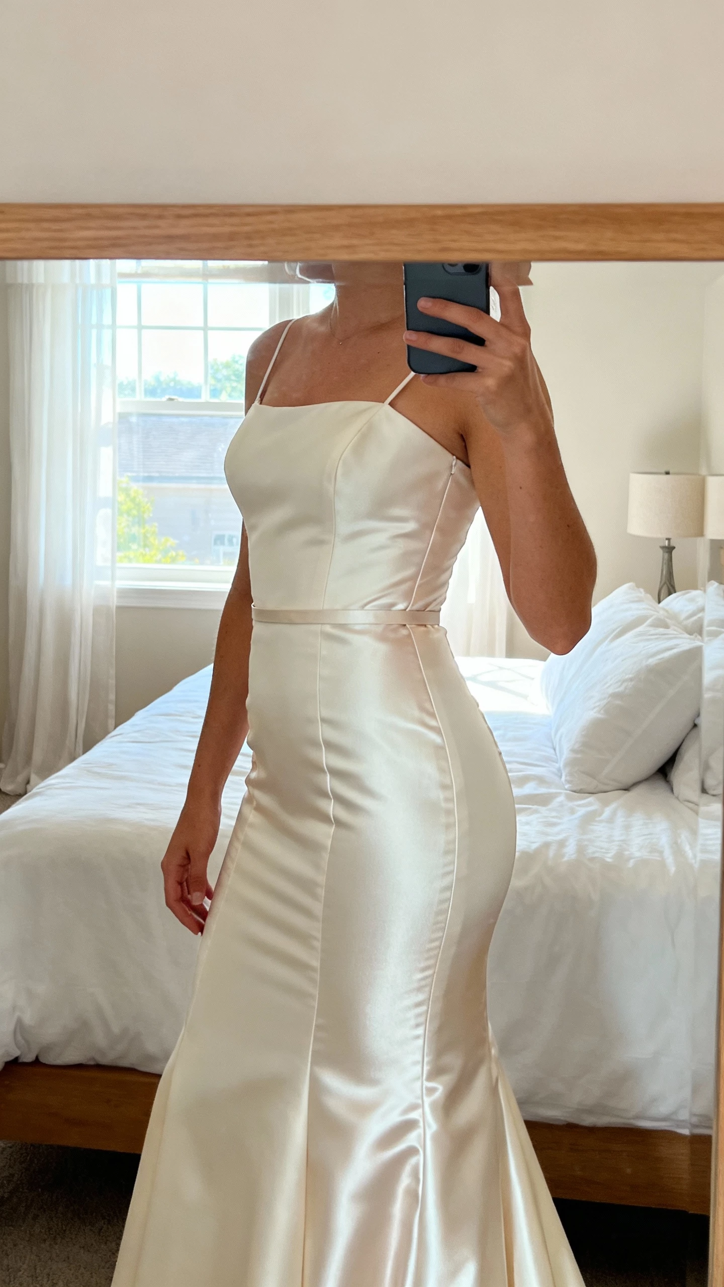 iPhone side mirror selfie of a woman in a bridal dress with refined seams and a silky lining that smooths the torso and hips; thin belt at waist, face not visible, bedroom mirror, afternoon window light, natural iPhone photo quality