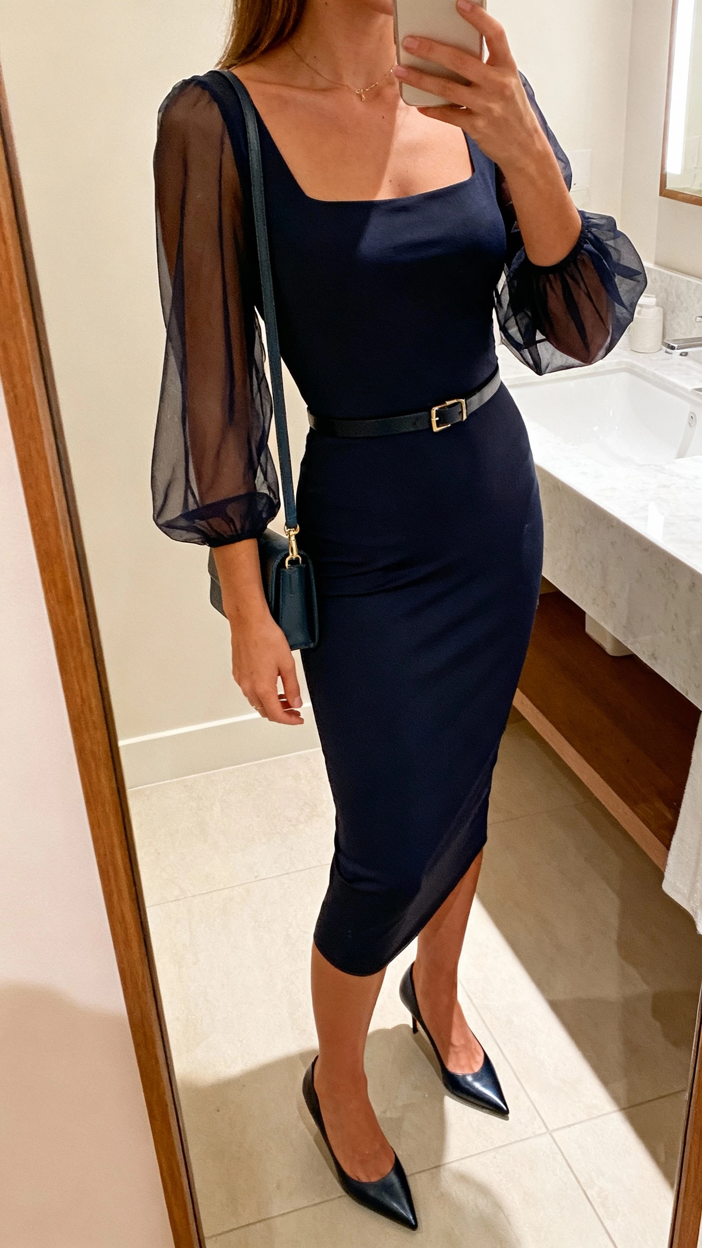 iPhone side-profile selfie cropped at neck of a woman wearing a bodycon midi dress with sheer sleeves, slim belt, pointed-toe pumps, small shoulder bag, face not visible, bathroom mirror with soft indoor light, natural iPhone photo quality.