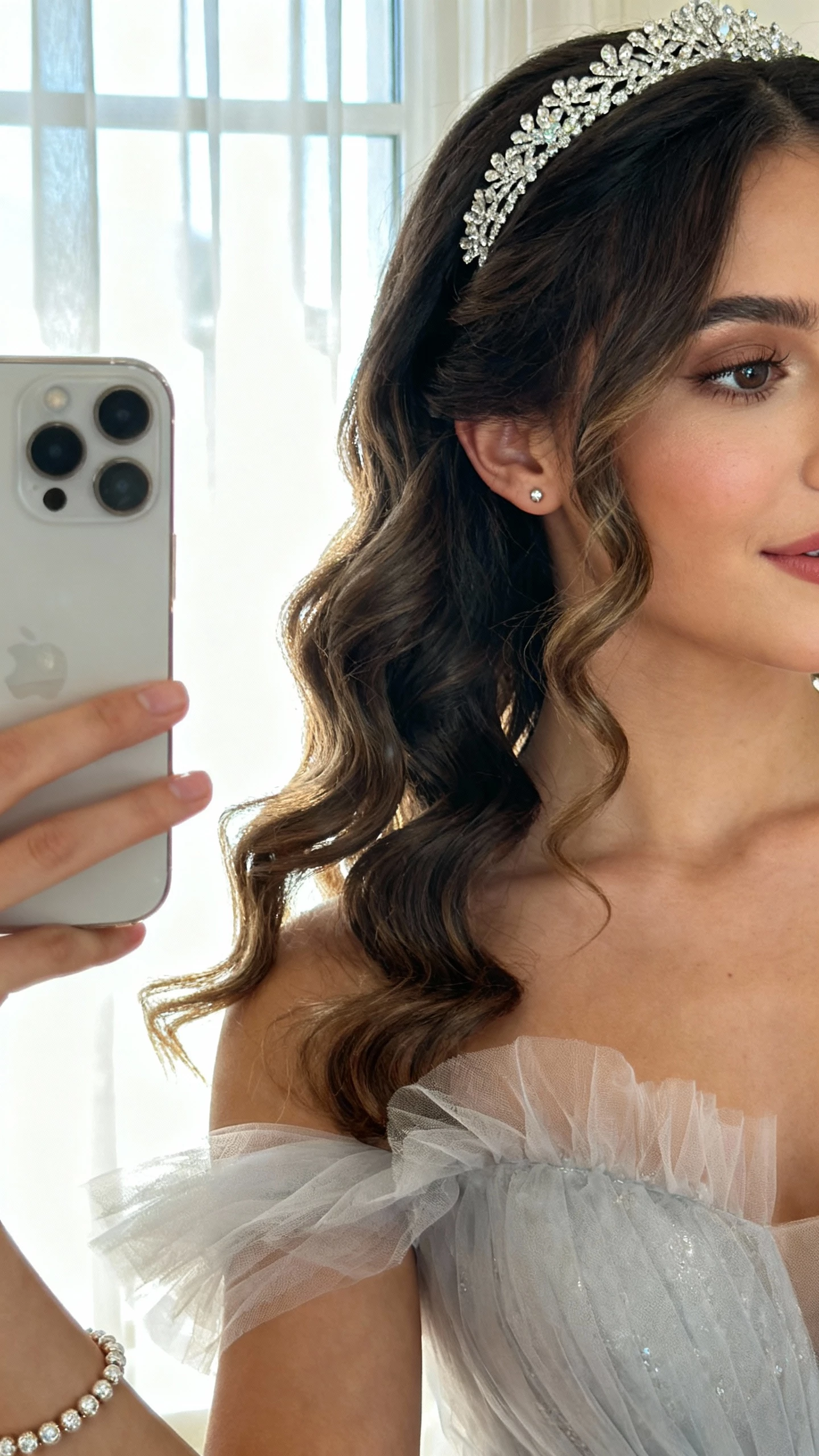 iPhone side-profile selfie cropped at shoulders showing cascading curls with a sparkling headband, princess-style tulle gown, delicate tennis bracelet, and subtle studs; face not visible, near window drapes, bright morning light, natural iPhone quality.