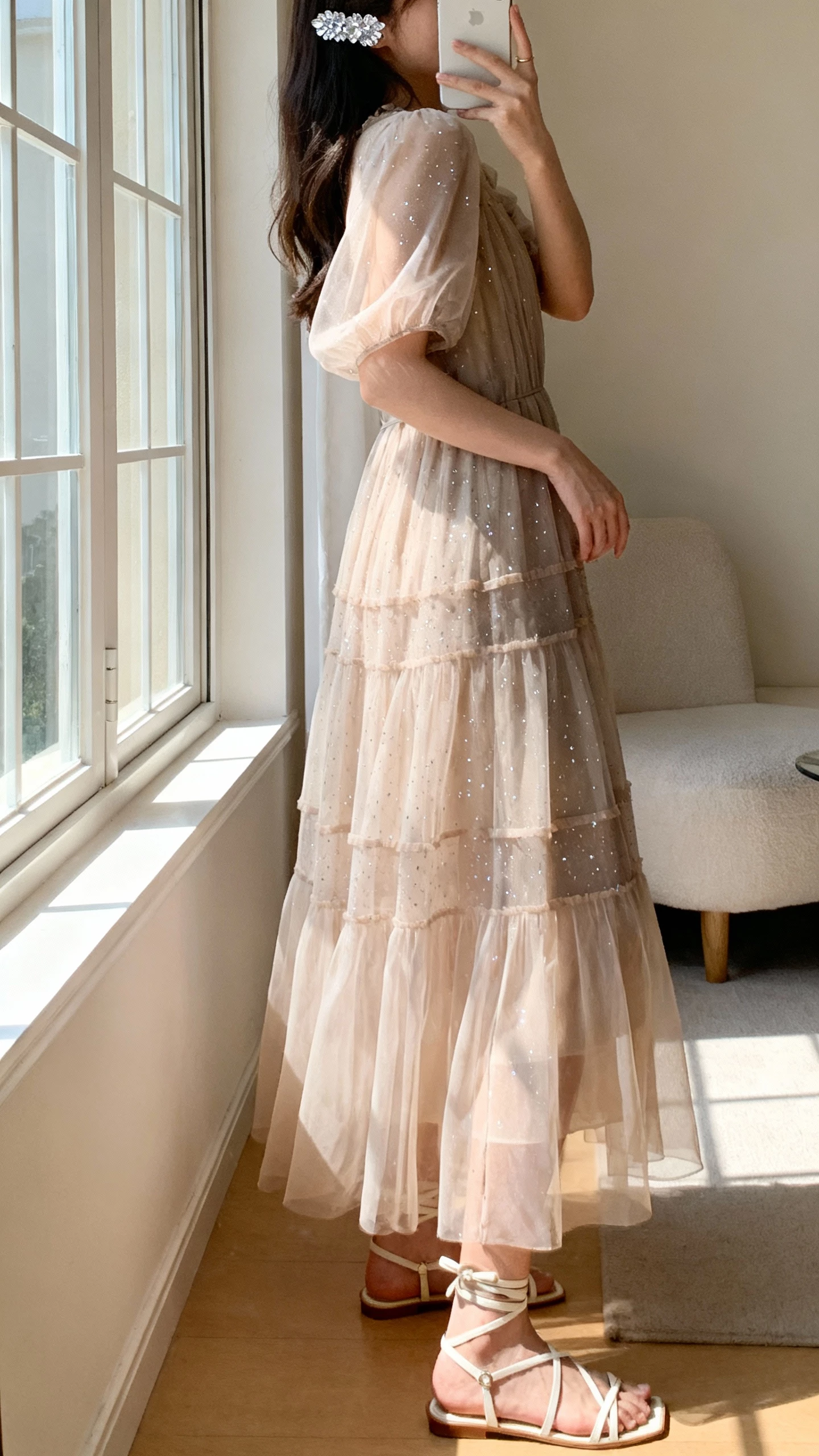 iPhone side-profile selfie of a woman in a romantic maxi dress with sheer layers and subtle sparkle, strappy sandals, crystal hair pin visible, face not visible, taken near a window in a softly lit living room, natural iPhone photo.