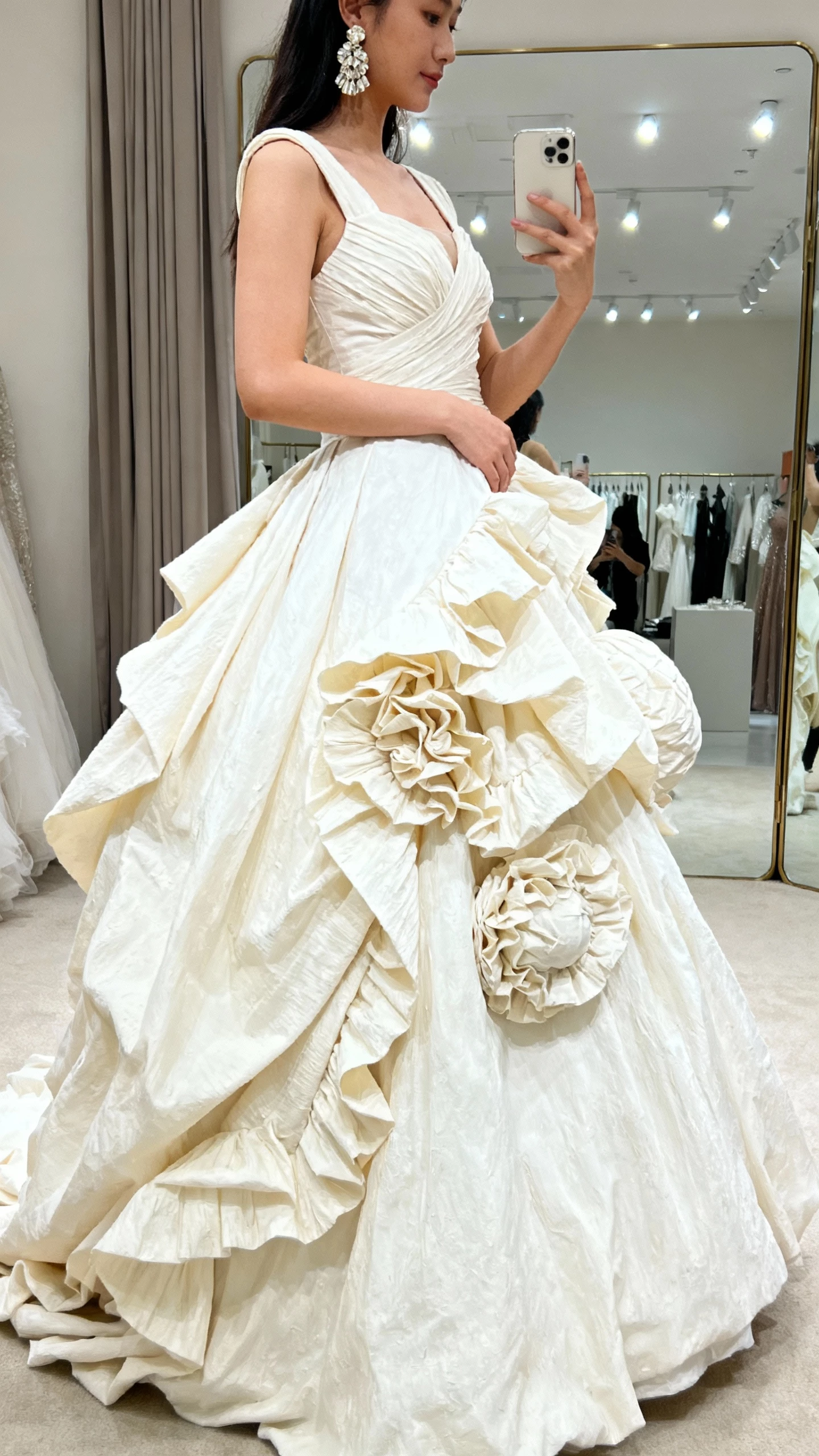 iPhone side-profile selfie of a woman wearing a sculptural ruffle ball gown with dramatic asymmetrical ruffles cascading down the skirt, structured bodice, and statement earrings, face not visible, boutique fitting room mirror, bright indoor lights, natural iPhone aesthetic