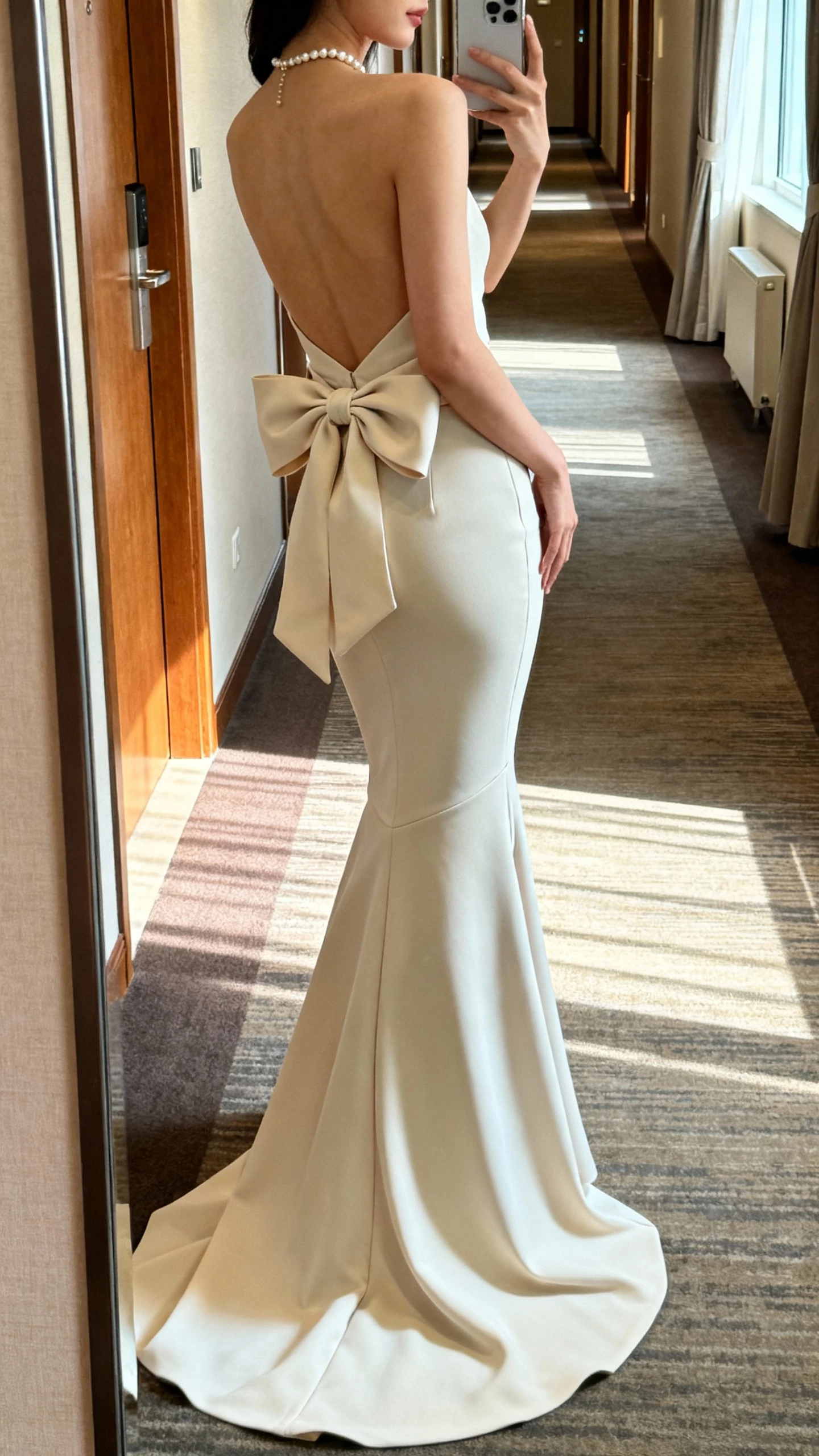 iPhone side-profile selfie of a woman wearing a sleek crepe mermaid gown with a low back and an oversized bow at the waist, clean train, petite pearl necklace, face not visible, hotel hallway mirror, natural morning light, iPhone photo aesthetic