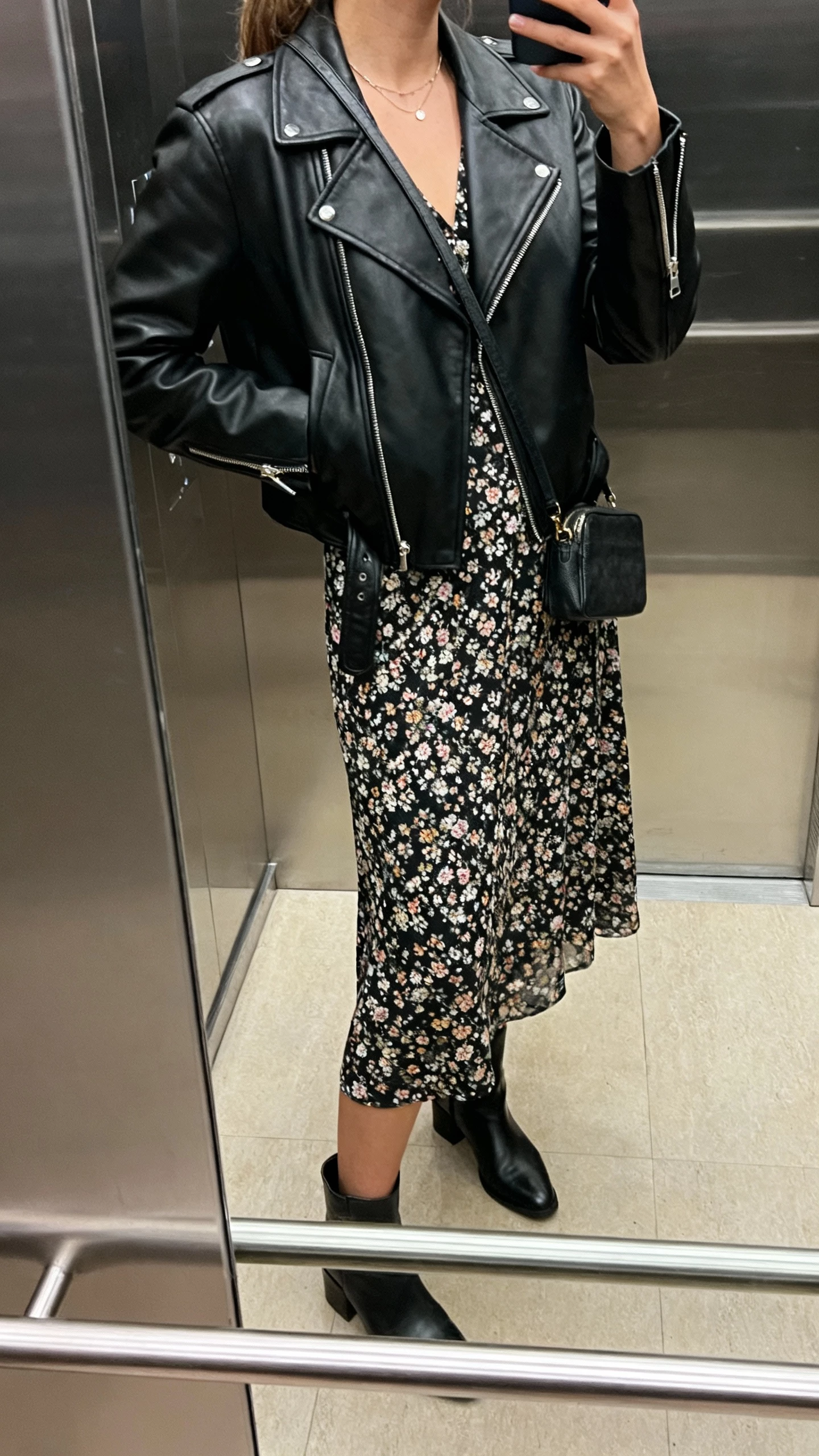 iPhone side-view mirror selfie of a woman in a black leather moto jacket over a floral midi dress, ankle boots, dainty necklace, small crossbody, face not visible, elevator mirror with indoor lighting, natural iPhone photo quality