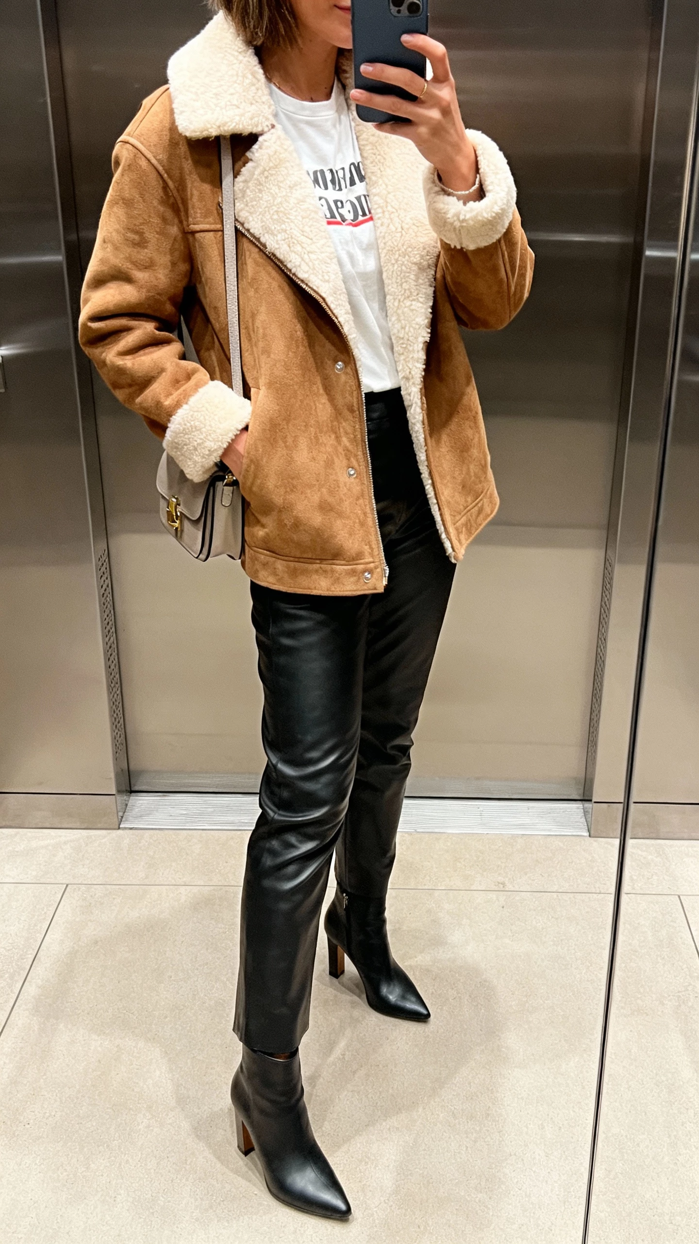 iPhone side-view mirror selfie of a woman in a faux shearling jacket over a tee, leather pants, heeled ankle boots, small shoulder bag, face not visible, elevator mirror, indoor lighting, casual iPhone photo.