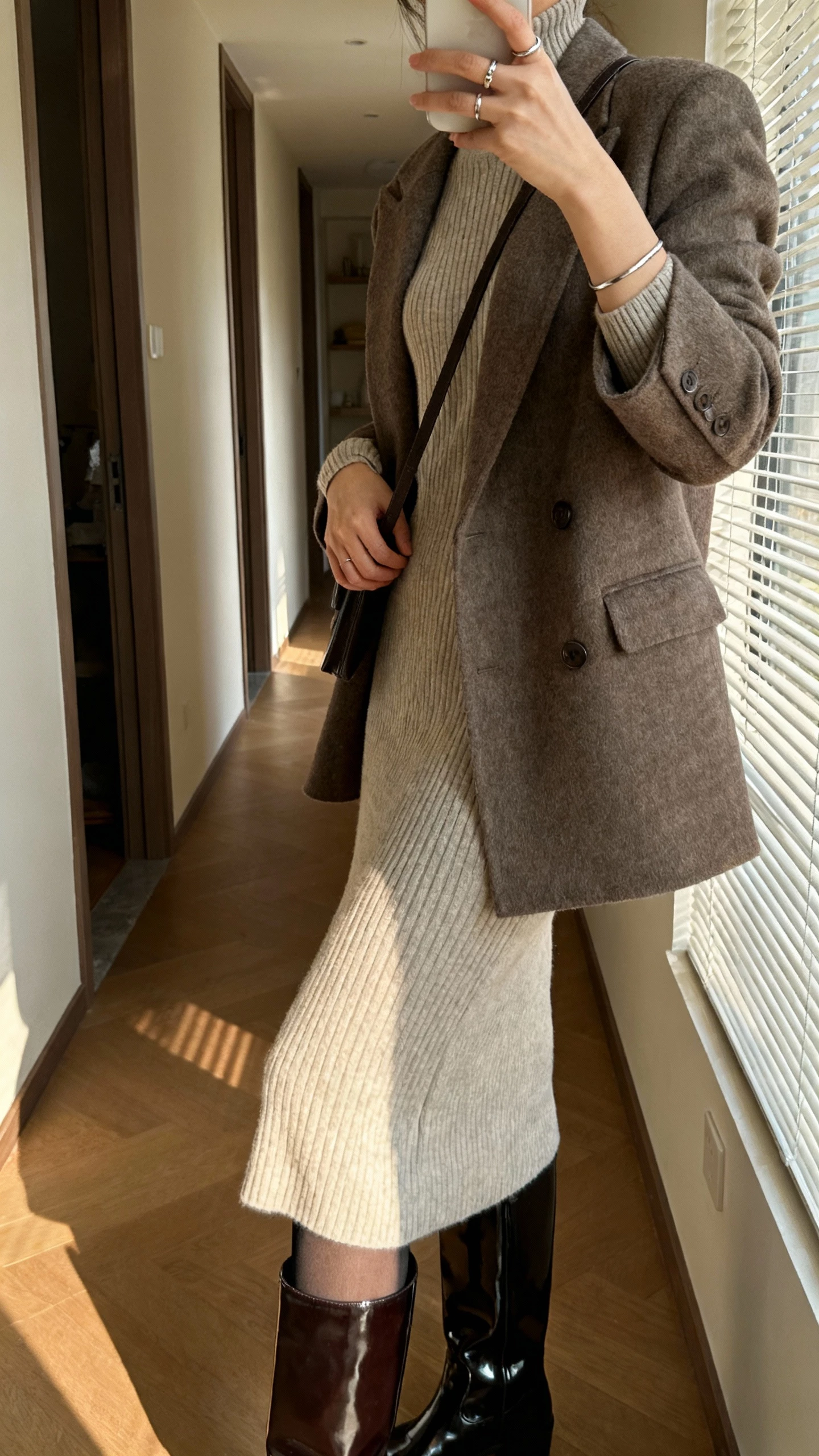iPhone side-view selfie cropped at shoulders of a woman in a long‑sleeve ribbed knit midi dress, patent‑leather knee‑high boots, single‑breasted tailored wool coat, minimal crossbody, fine stacking rings and slim cuff, opaque tights; apartment hallway, face not visible, afternoon window light, natural iPhone photo quality.