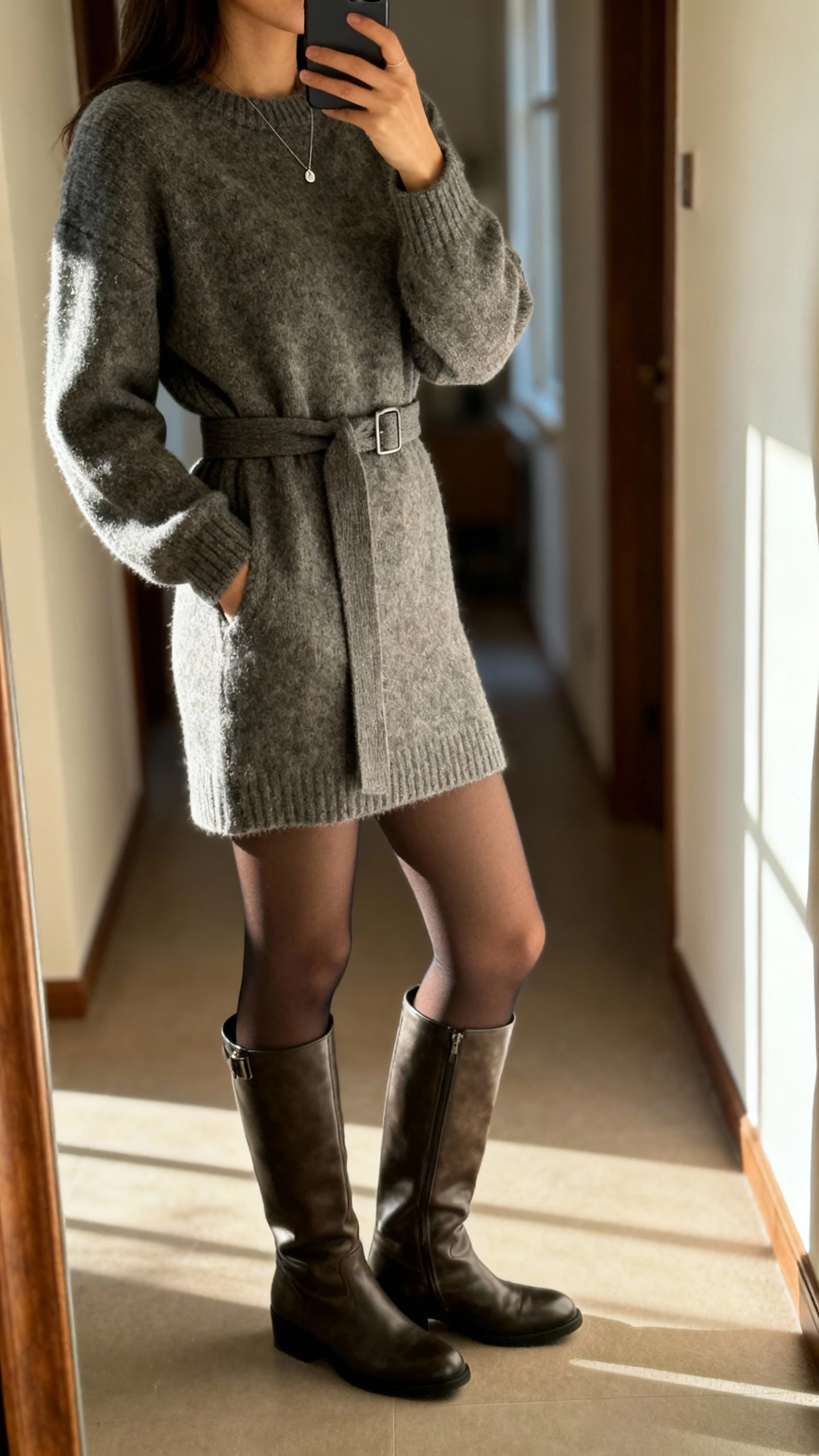 iPhone side-view selfie of a woman in a belted knit dress with tall boots, opaque tights, minimal pendant, face not visible, hallway mirror, afternoon window light, casual iPhone photo.