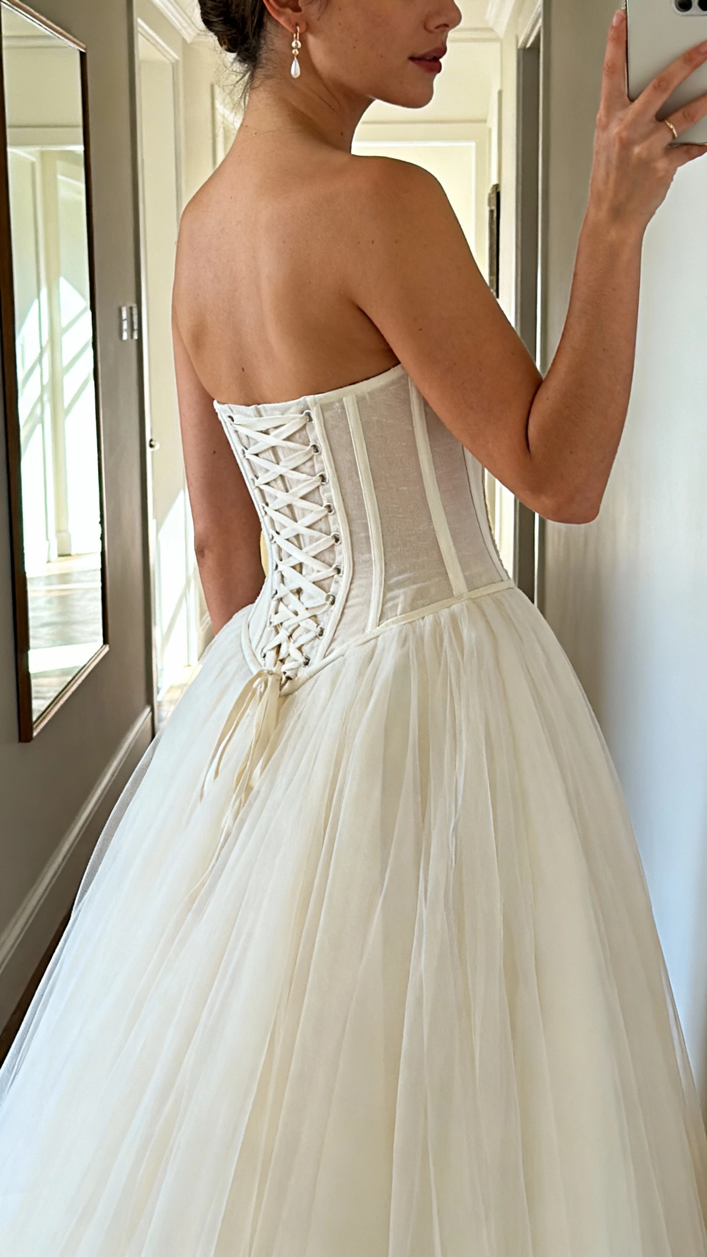 iPhone side-view selfie of a woman in a corset-bodice A-line wedding dress with visible boning, lace-up back, layered tulle skirt, and delicate earrings; face not visible, hallway mirror with natural daylight, natural iPhone photo quality.