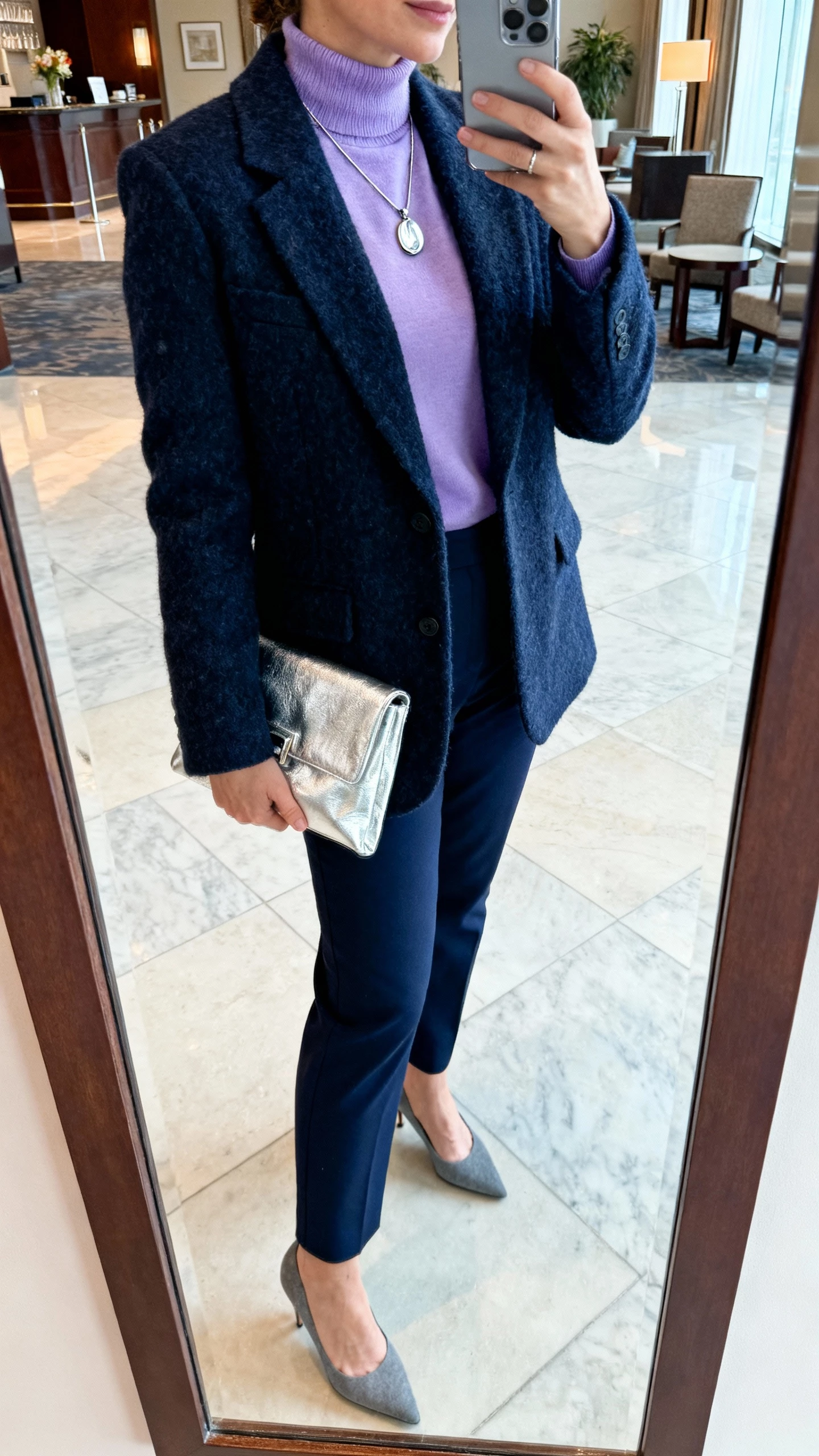 iPhone side-view selfie of a woman in a lavender mock-neck top, navy wool blazer, silver pendant necklace, navy trousers, silver clutch, gray heels, face not visible, hotel lobby mirror with cool indoor lighting, casual iPhone aesthetic