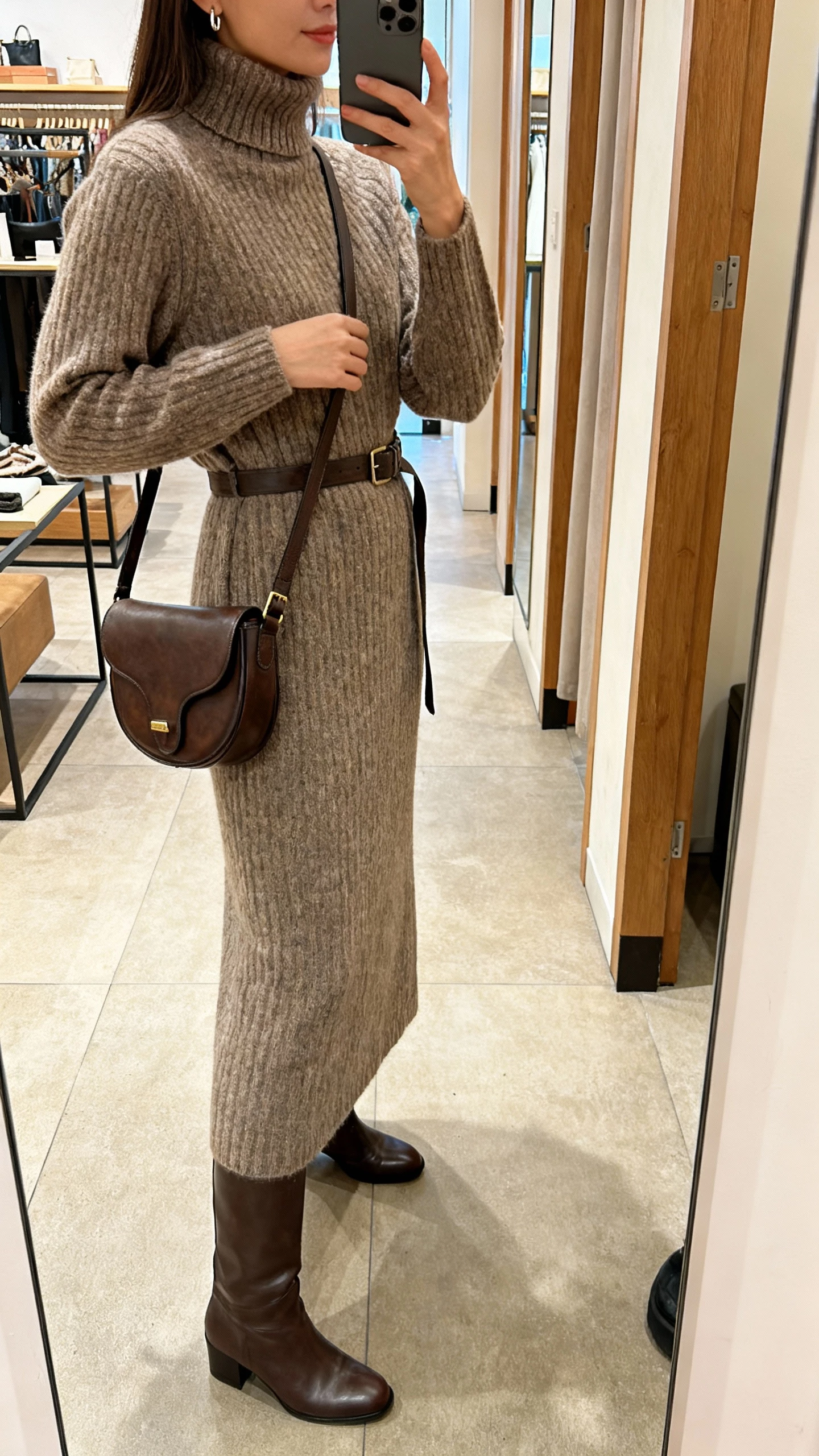 iPhone side-view selfie of a woman in a long knit turtleneck dress cinched with a belt, knee-high boots, crossbody saddle bag, minimal earrings, face not visible, boutique fitting-room mirror, warm indoor lighting, natural iPhone photo quality.