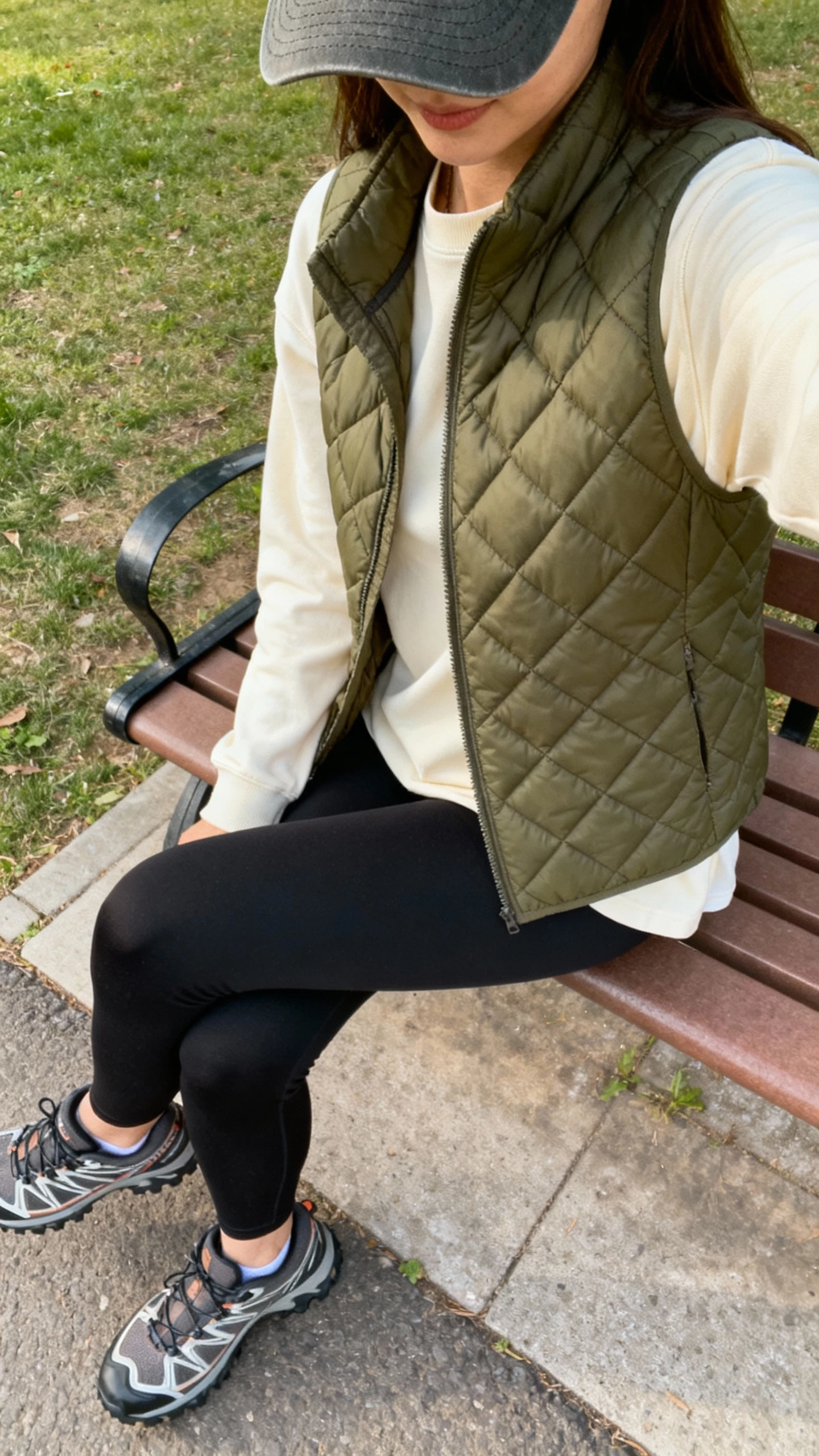 iPhone side-view selfie of a woman in a quilted olive vest over a cream long-sleeve tee, black leggings, trail sneakers, baseball cap, face not visible, park bench setting, natural daylight, iPhone photo quality