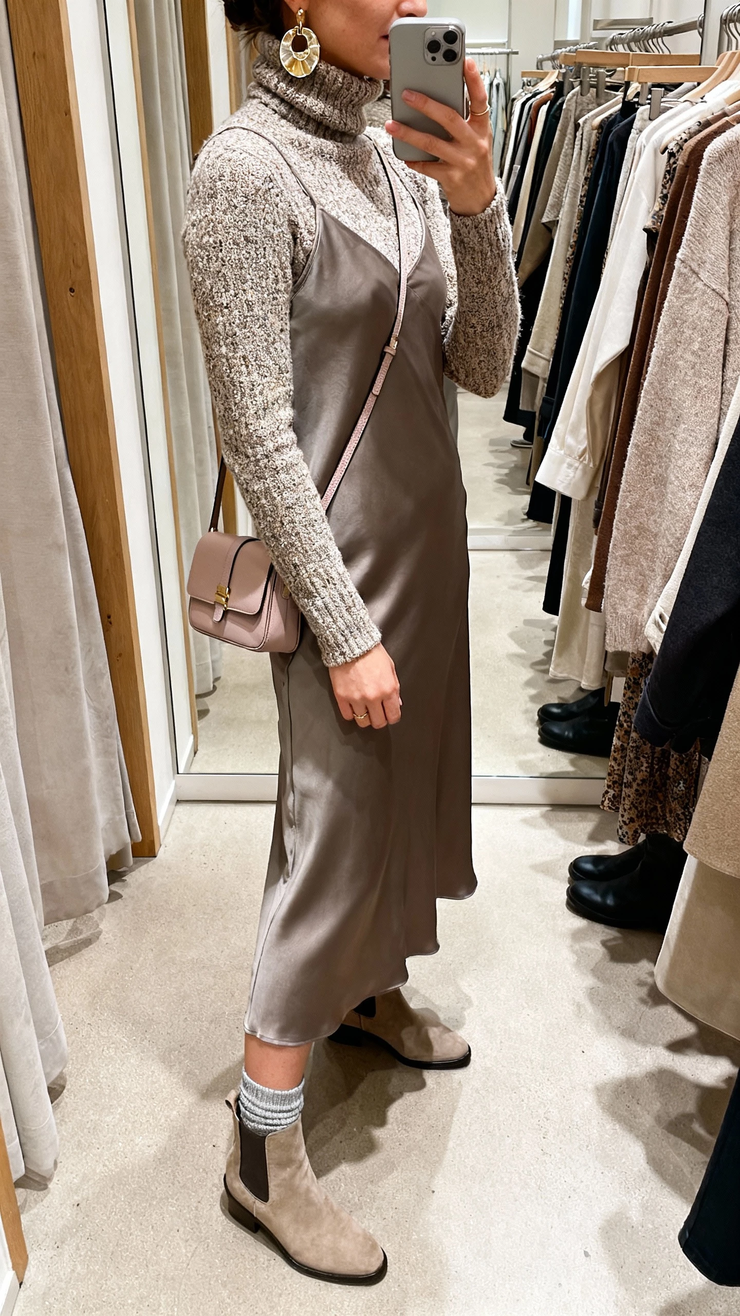 iPhone side-view selfie of a woman layering a textured turtleneck under a midi slip dress with sock boots, statement earrings and a mini shoulder bag, face not visible, boutique dressing room, soft indoor lighting, casual iPhone aesthetic