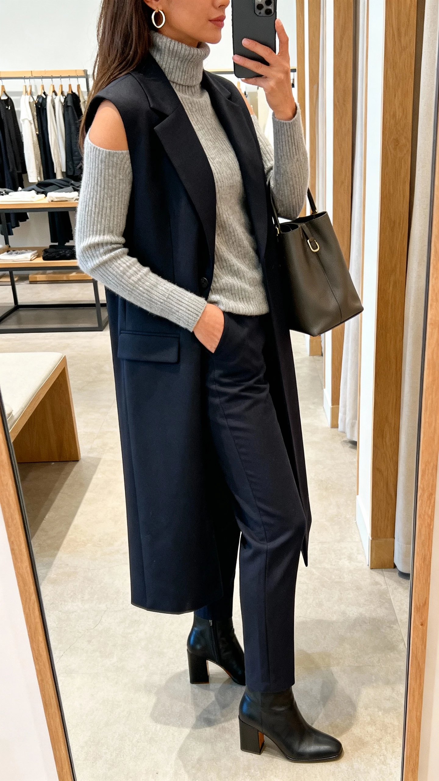 iPhone side-view selfie of a woman wearing a fitted turtleneck under a sleeveless sweater and a long tailored vest, with slim trousers and block-heel boots, minimal hoops and a structured bag, face not visible, boutique fitting room mirror, indoor lighting, natural iPhone photo quality.