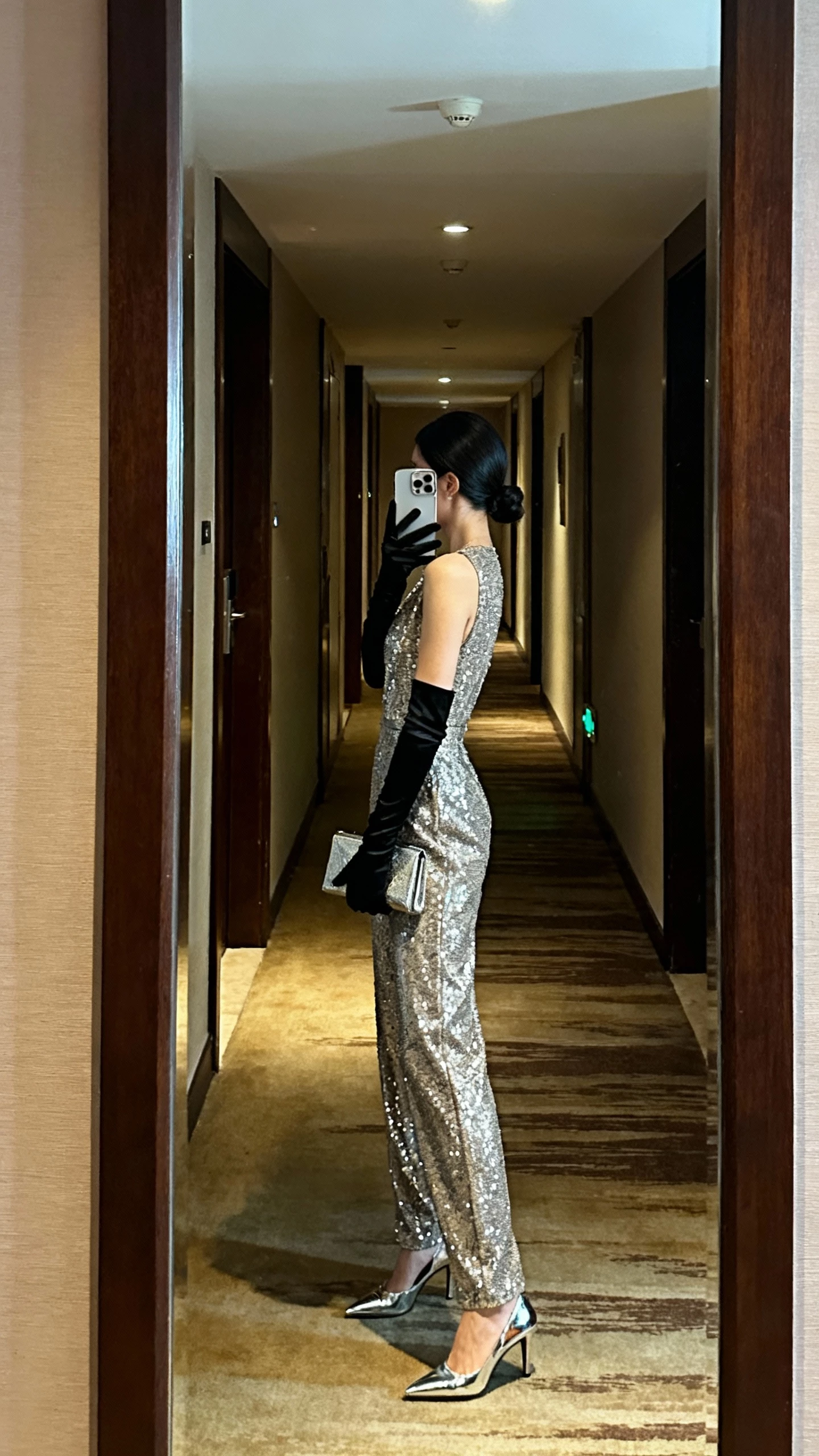 iPhone side-view selfie of a woman wearing a glam jumpsuit with opera gloves and sleek hair, metallic heels, and a clutch, face not visible, hotel corridor mirror, soft evening lighting, casual iPhone photo.