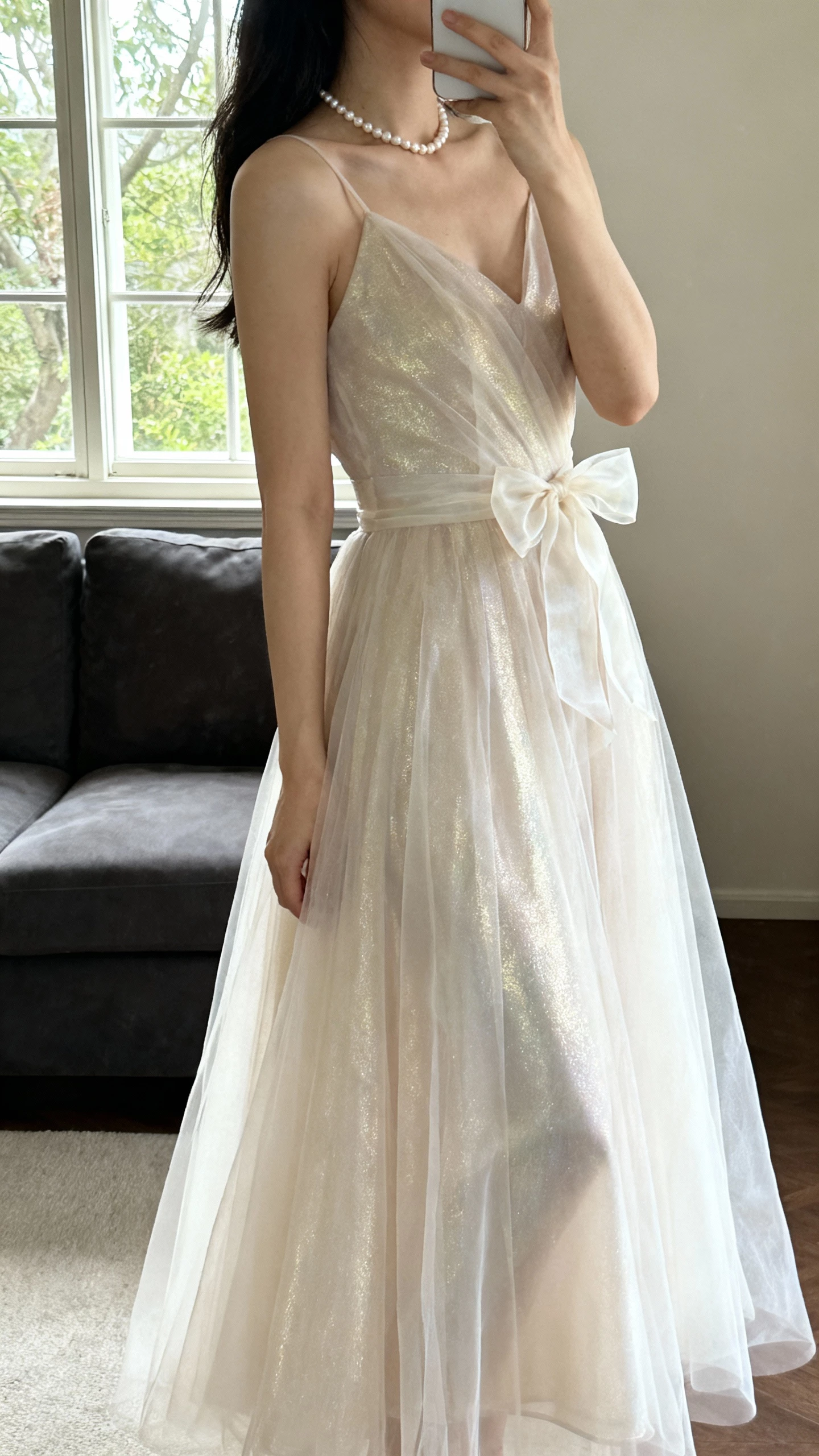 iPhone side-view selfie of a woman wearing a luxe organza bridal gown with light sheen, airy A-line skirt, organza bow at waist, simple pearl necklace, face not visible, living room near window, gentle daylight, natural iPhone photo quality