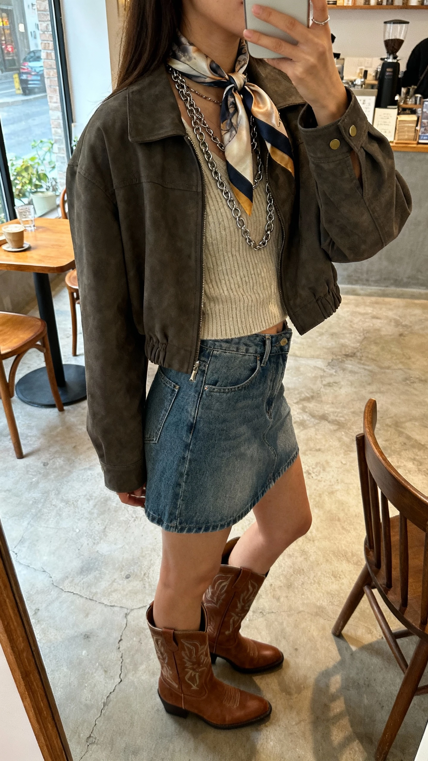 iPhone side-view selfie of a woman wearing a silk scarf tied at the neck, Western boots, layered chains over a simple knit, denim skirt, and a cropped jacket, face not visible, artsy cafe setting with natural daylight, natural iPhone photo quality.