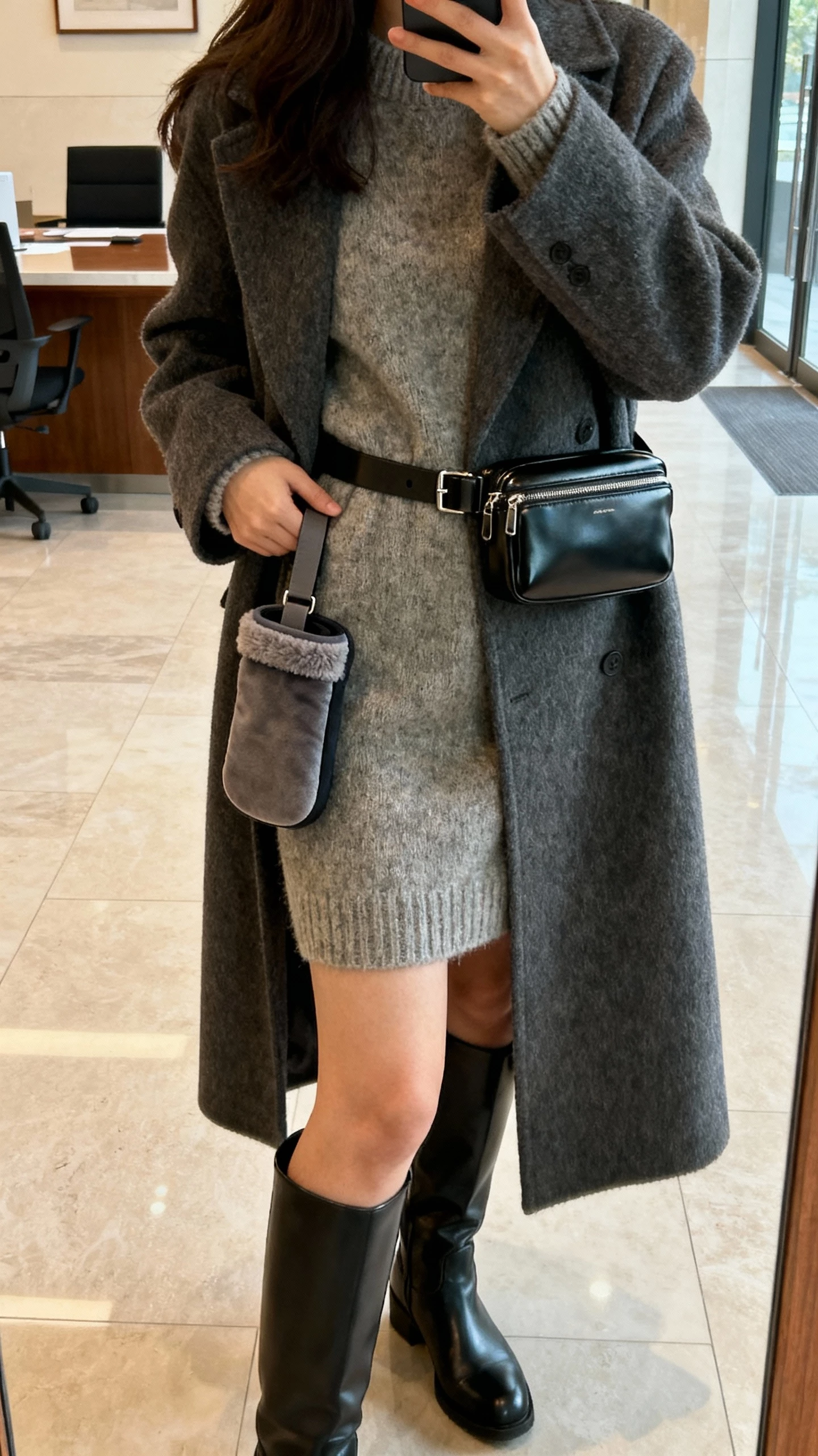 iPhone side-view selfie of a woman wearing a sleek belt bag at the waist and a hand warmer (muff) slung with a strap, long wool coat, sweater dress, knee-high boots, face not visible, office lobby mirror, soft indoor lighting, natural iPhone photo.