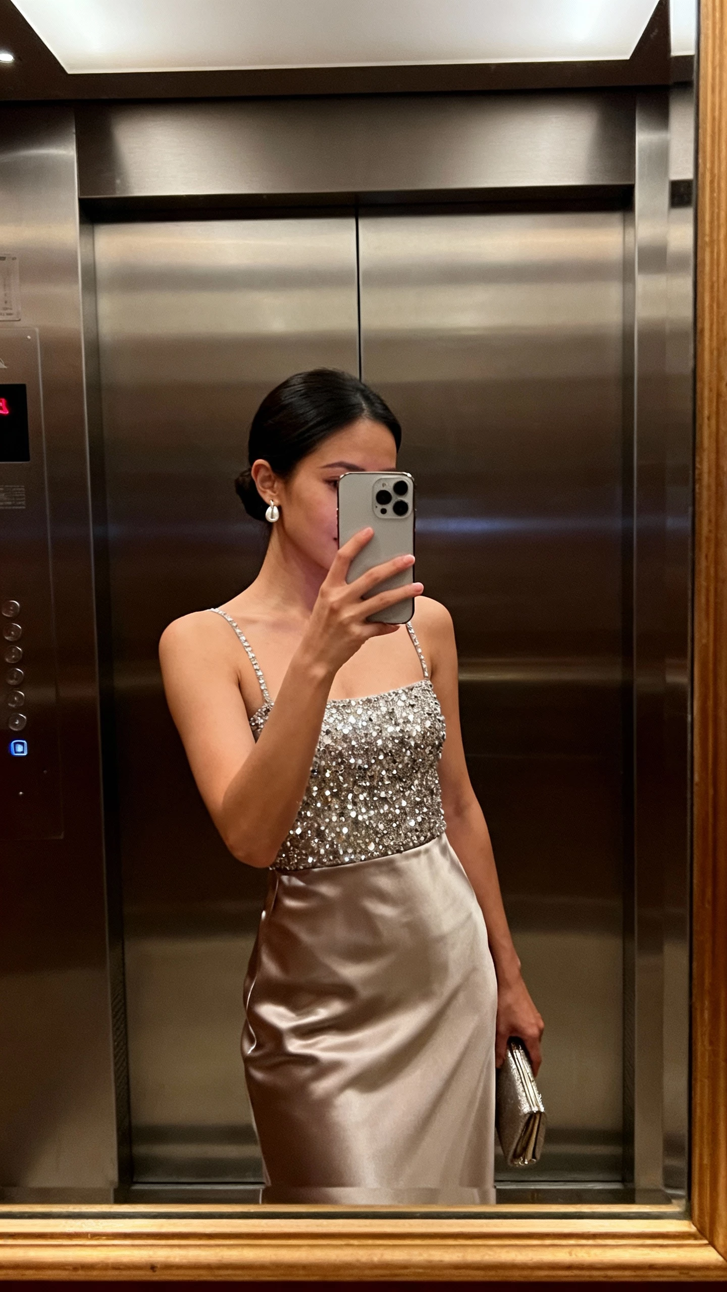iPhone side-view selfie of a woman wearing a sparkle-embellished bodice with a smooth satin skirt, simple earrings and clutch, face not visible, elevator mirror, evening indoor lighting, natural iPhone photo quality