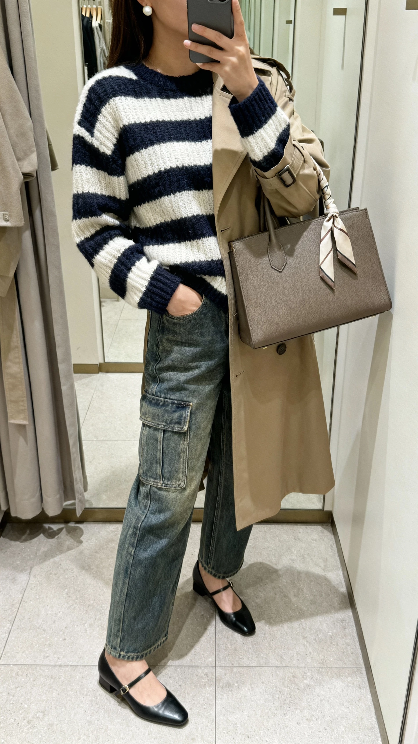 iPhone side-view selfie of a woman wearing a striped knit sweater with straight-leg mid-wash cargo jeans, slingback flats, trench coat, structured satchel with a thin scarf tied to the handle, simple pearl studs, face not visible, boutique fitting-room mirror, soft natural lighting, casual iPhone photo.