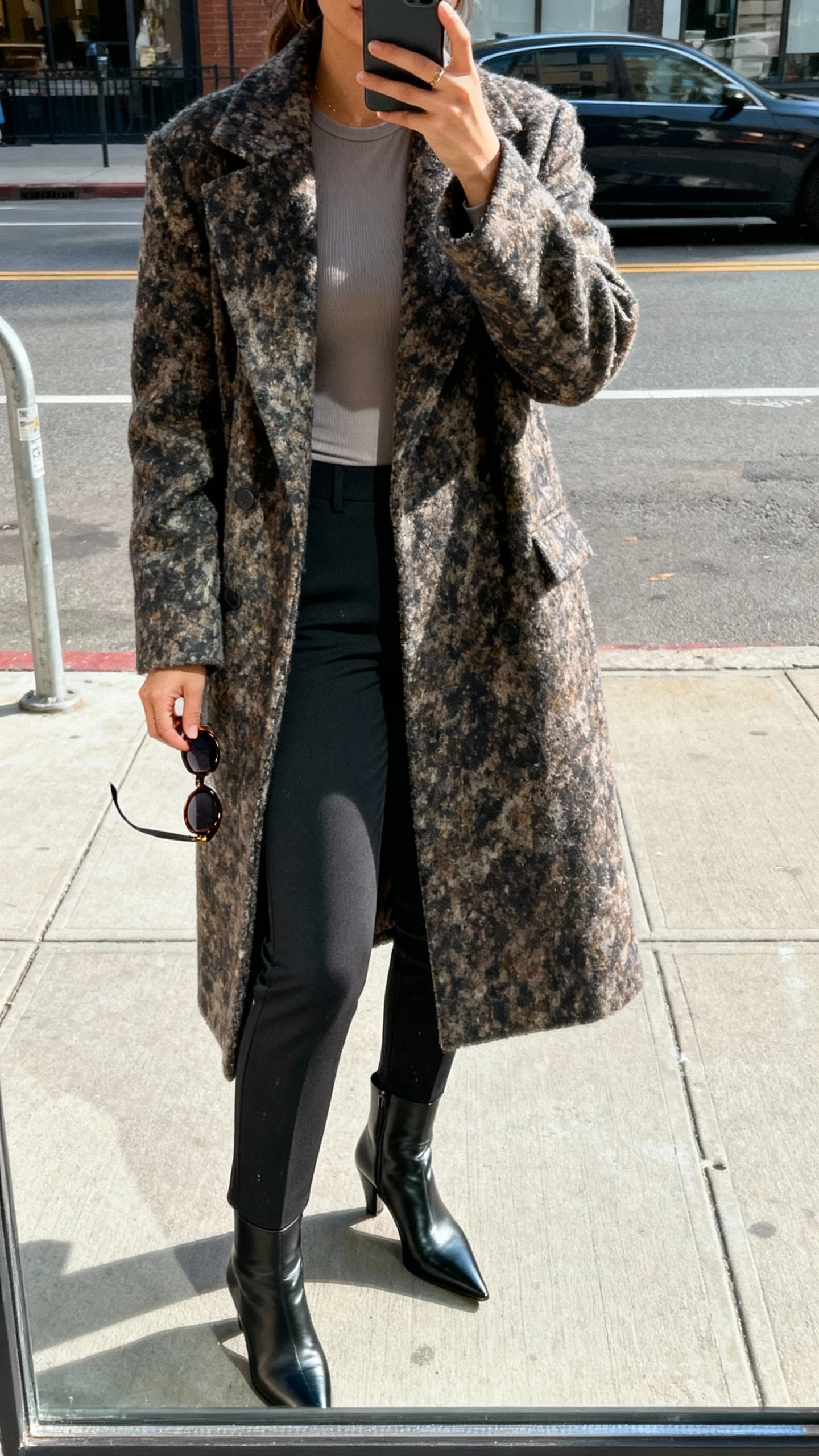 iPhone sidewalk mirror-window selfie of a woman wearing a statement coat over a minimal base (simple fitted top and slim trousers), pointed boots, and sunglasses in hand, face not visible, city street with natural daylight, natural iPhone photo quality