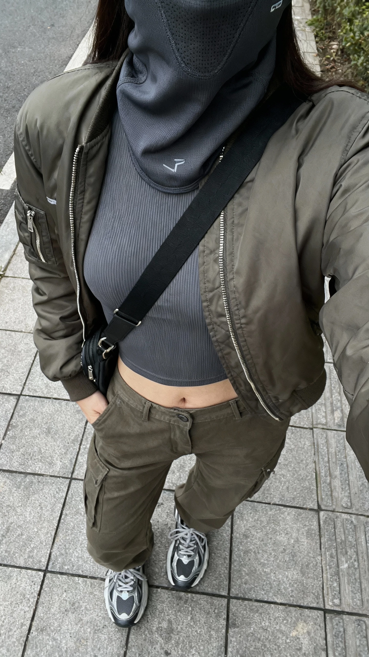 iPhone sidewalk selfie from chest down showing a woman in thermal base layers under a bomber jacket with a sleek balaclava, cargo pants, chunky sneakers, crossbody, face not visible, overcast natural daylight, casual iPhone look.