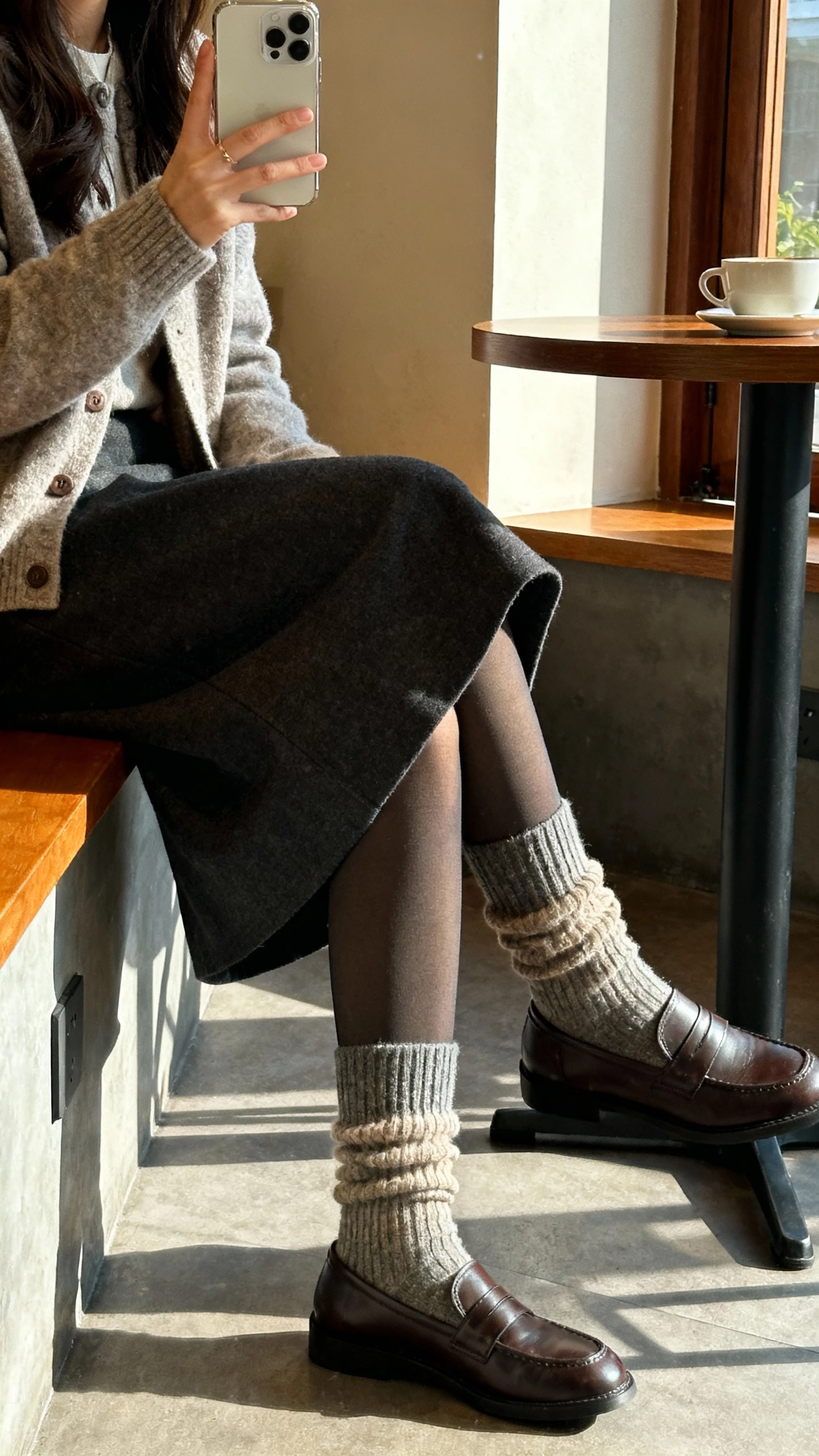 iPhone sitting selfie cropped at knees of a woman in cozy wool socks layered over opaque tights, midi skirt, cardigan, loafers, face not visible, cafe corner with morning light, natural iPhone photo quality.