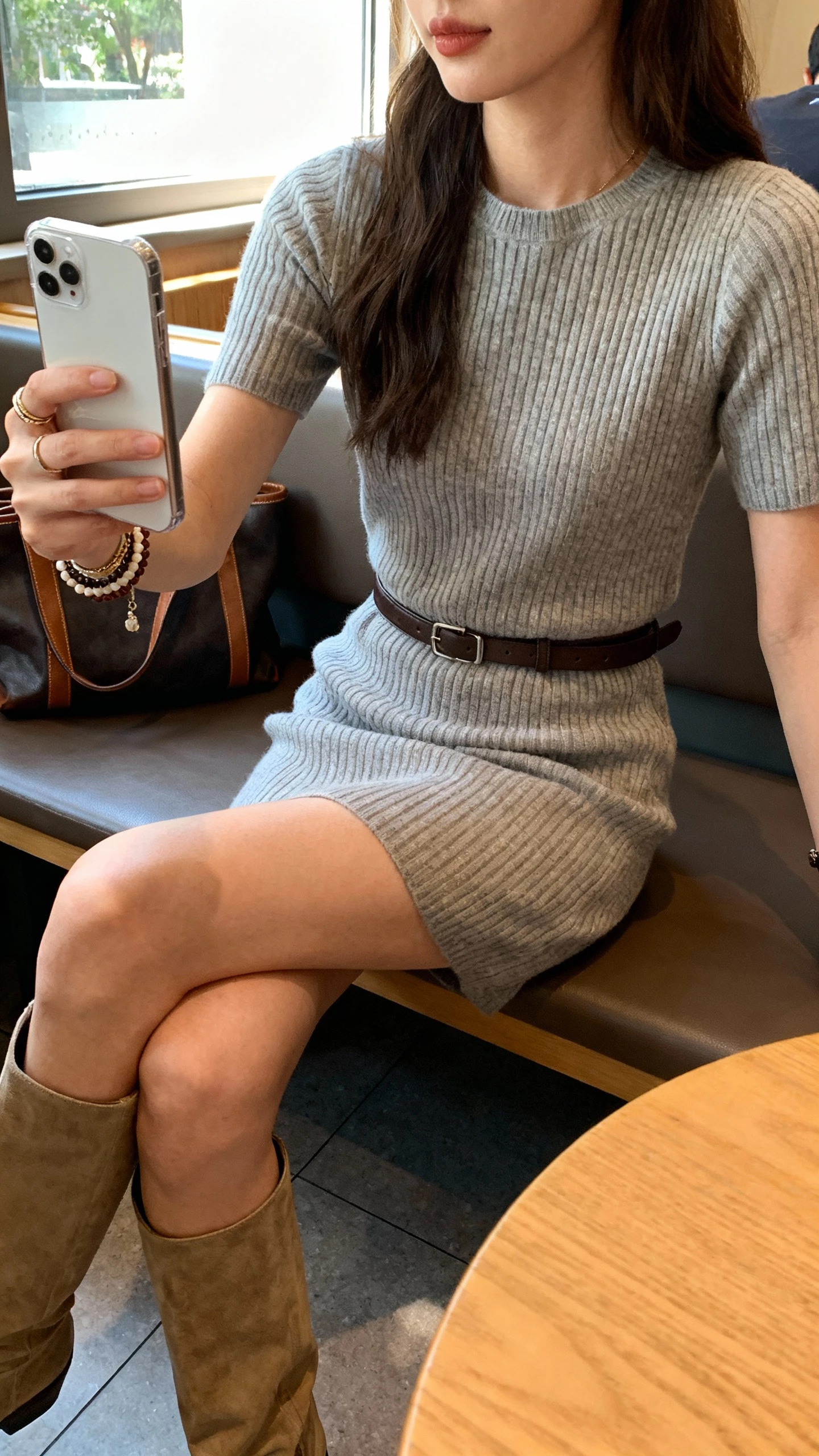 iPhone sitting selfie from cafe bench of a woman in a ribbed knit dress with a waist belt, tall boots, layered bracelets, and a tote on the side, face not visible, cropped at neck, cozy cafe setting with window light, natural iPhone photo quality.
