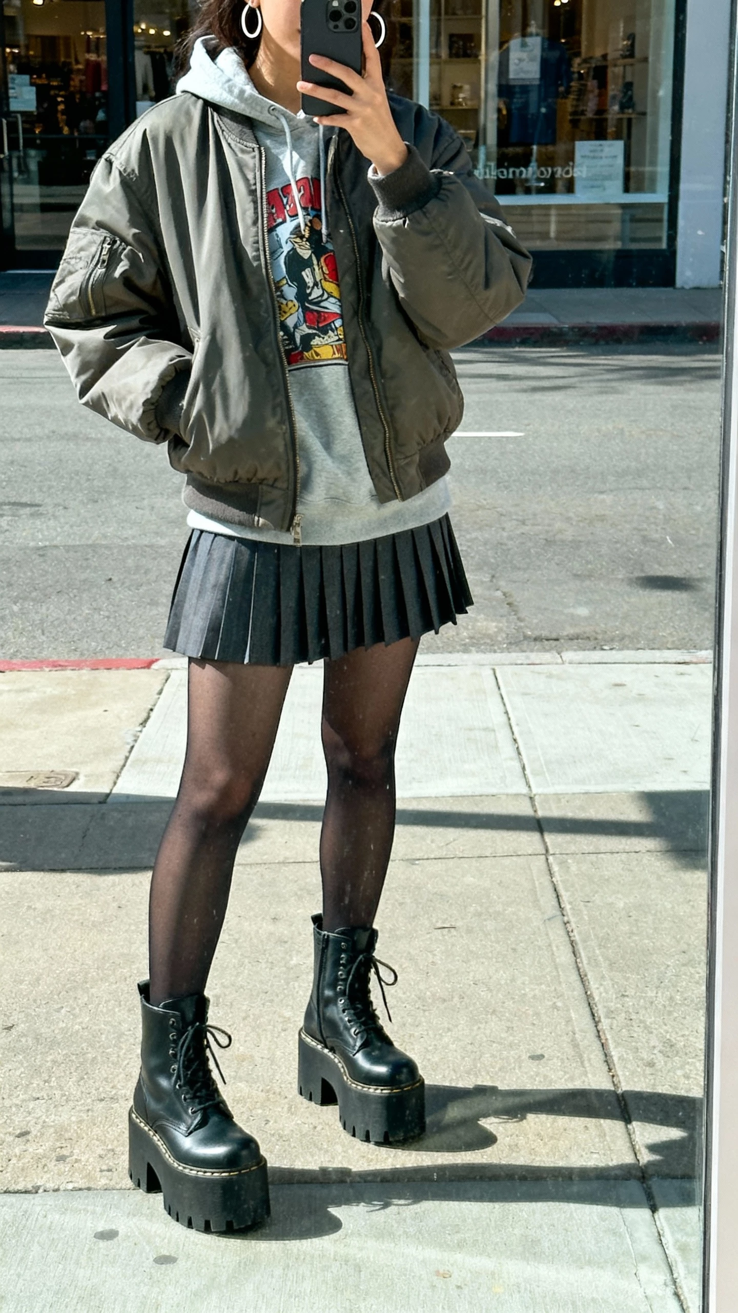 iPhone street mirror reflection selfie (storefront glass) of a woman in platform combat boots, oversized bomber jacket, graphic hoodie, pleated mini skirt with tights, and hoop earrings, face not visible, urban sidewalk, daylight, casual iPhone photo.