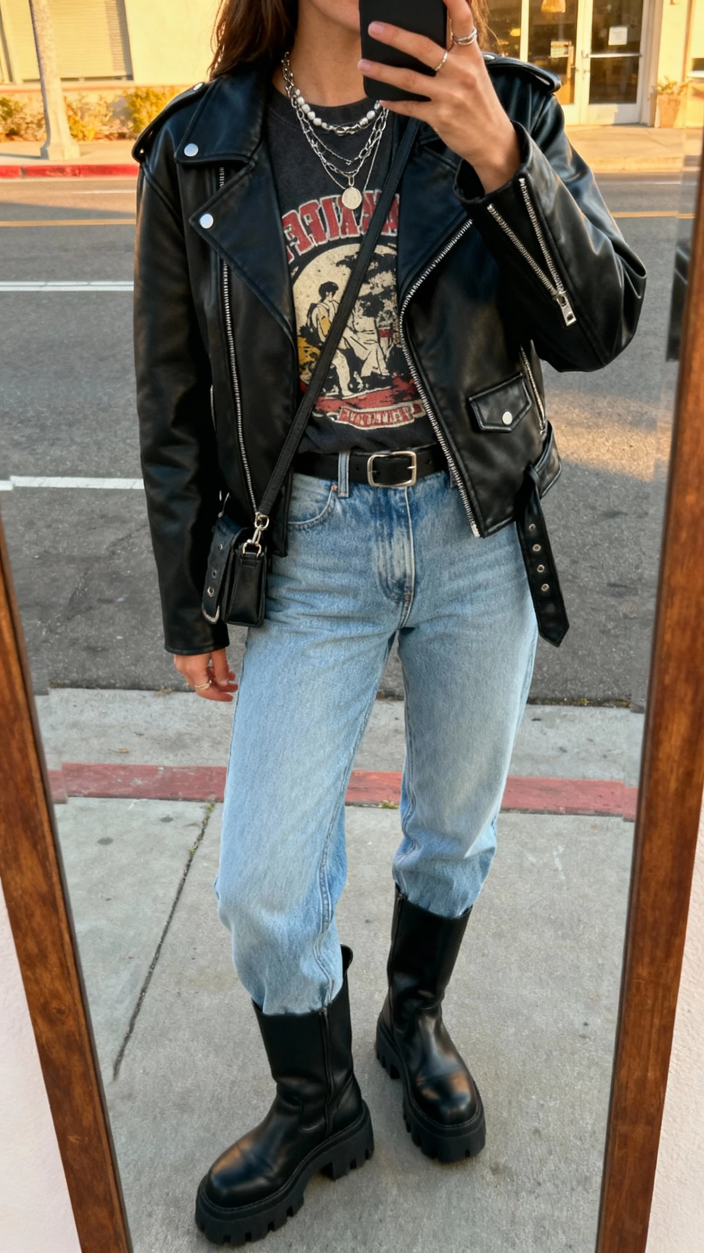 iPhone street mirror selfie of a woman in a vintage graphic tee tucked into light blue jeans, black leather moto jacket, chunky black boots, layered necklaces, small crossbody, face not visible, late afternoon natural light, natural iPhone aesthetic