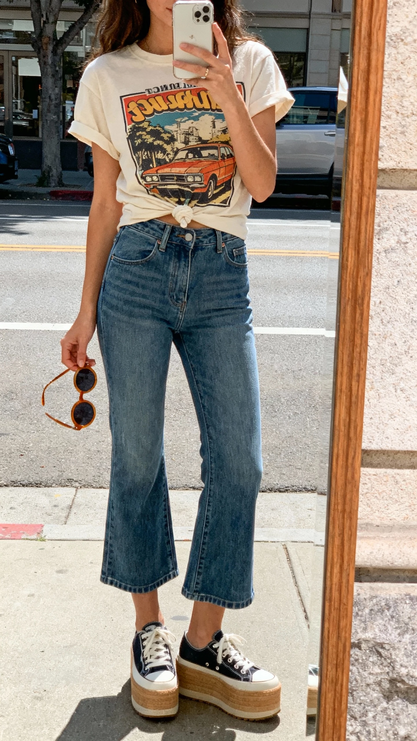 iPhone street mirror selfie showing a woman in a vintage graphic tee tucked into cropped flare jeans, platform sneakers, round sunglasses in hand, face not visible, bright daylight, casual iPhone photo.