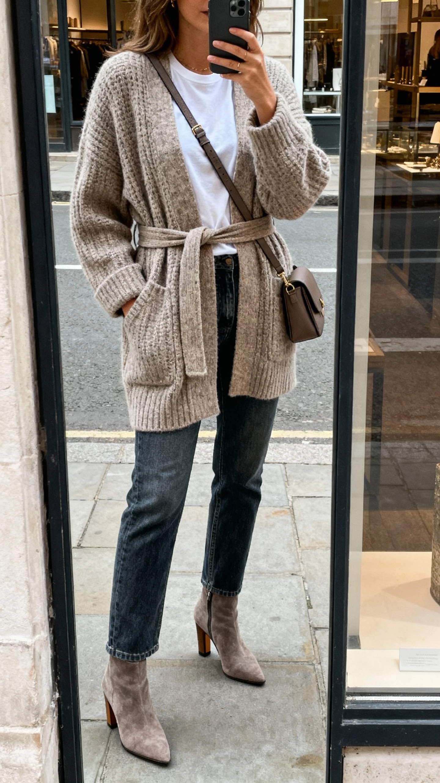 iPhone street-mirror (shop window) selfie of a woman in a belted knit coatigan over a tee, straight jeans, heeled booties, and a crossbody bag, face not visible, overcast daylight, casual iPhone aesthetic.