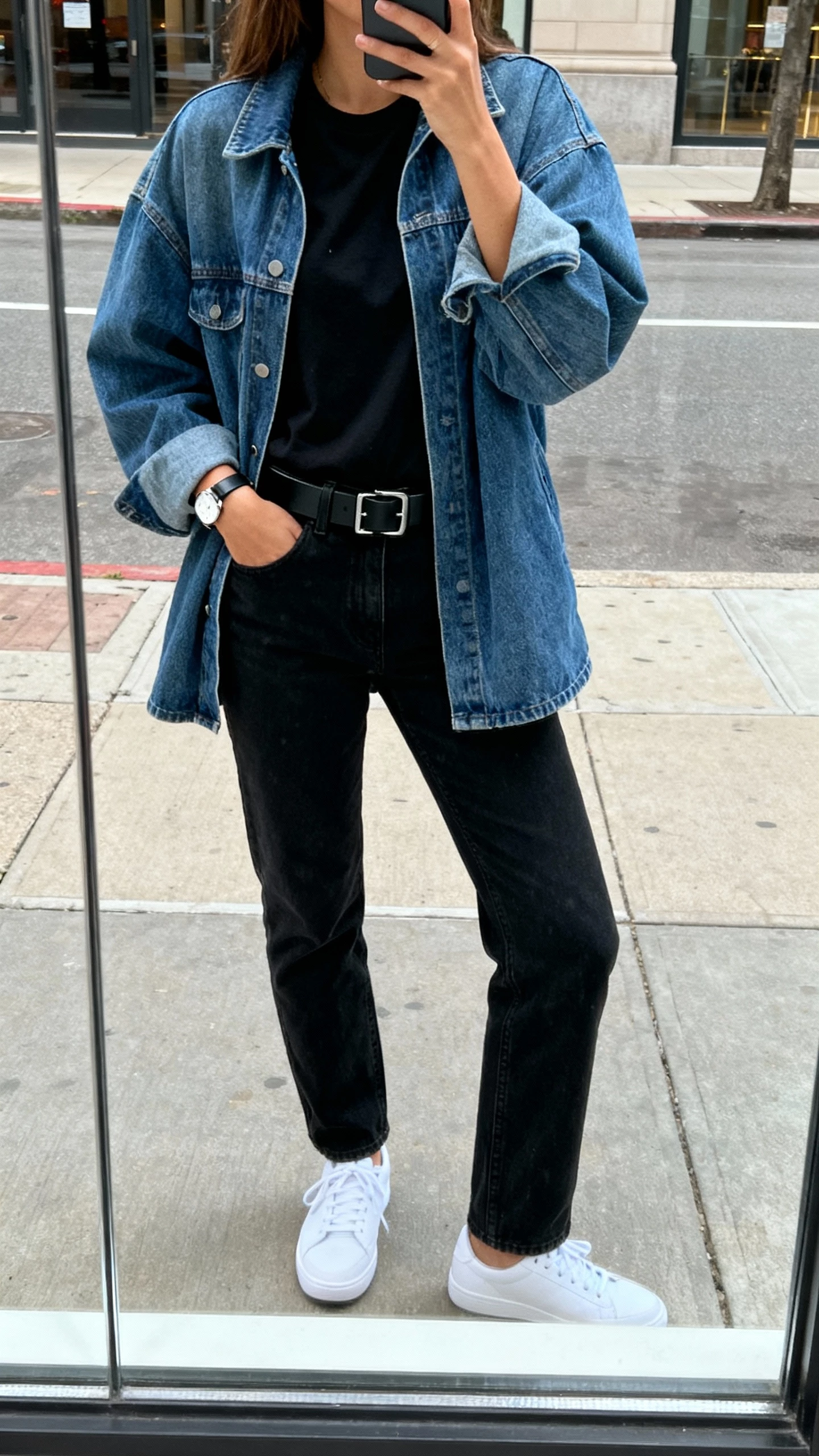 iPhone street mirror window selfie of a woman in an oversized denim jacket over an all-black base (black tee and black jeans), white sneakers, black belt and simple watch, face not visible, city sidewalk reflection, natural daylight, natural iPhone photo quality