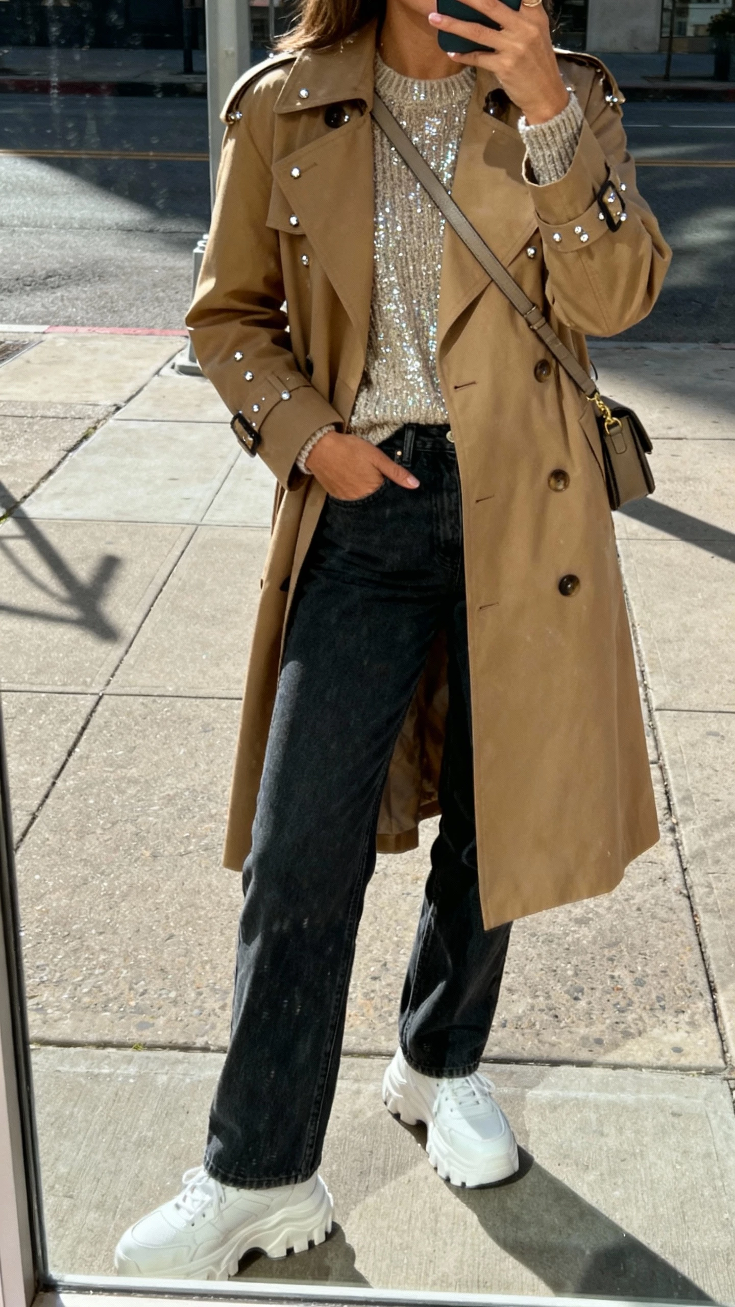 iPhone street mirror-window selfie of a woman wearing a classic tan trench coat over a shimmer knit top, straight dark jeans, chunky white sneakers, crossbody bag, and subtle sparkly studs, face not visible, natural daylight on a city sidewalk, casual iPhone photo quality