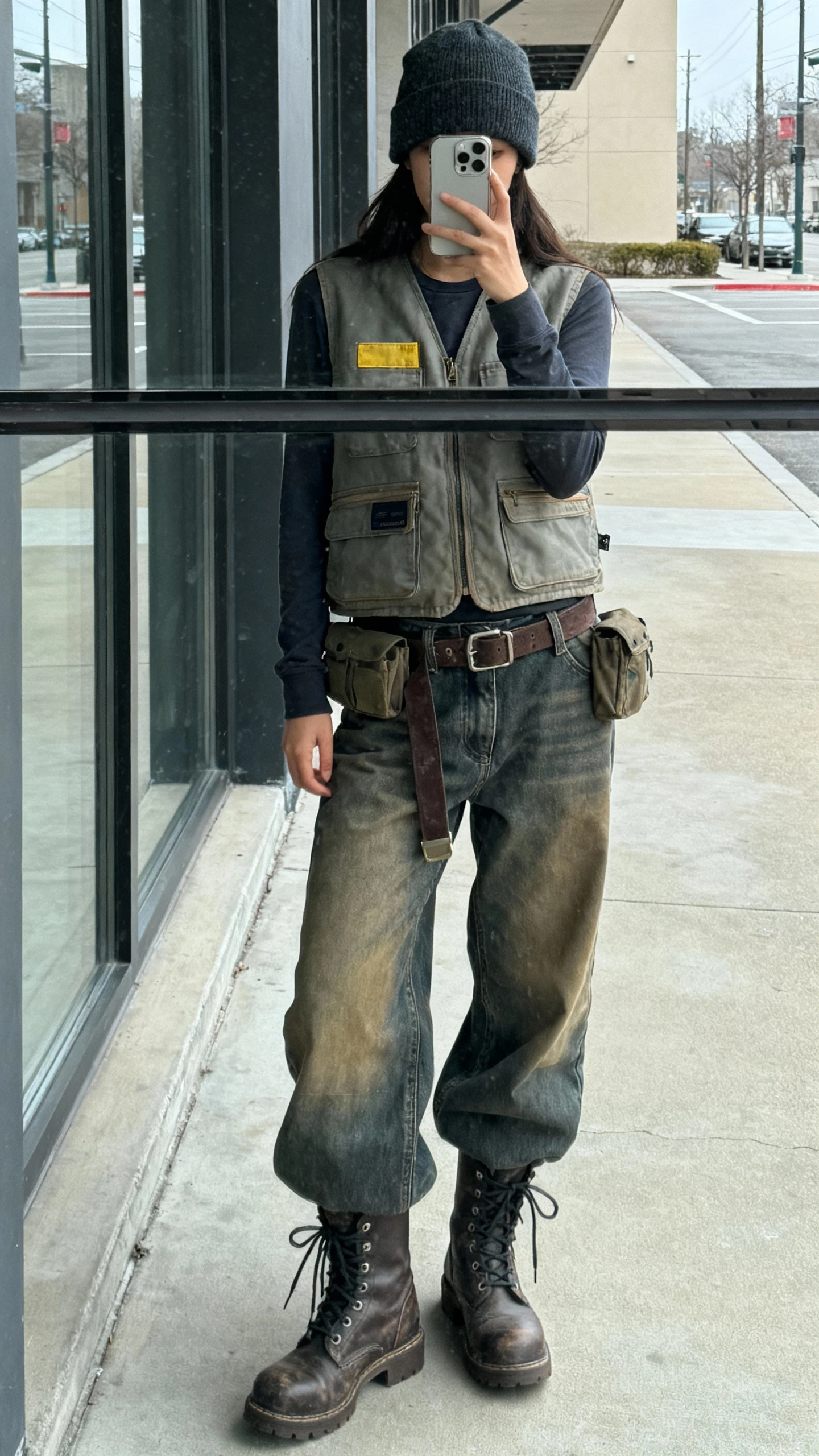 iPhone street mirror window selfie of a woman wearing a workwear vest over a long-sleeve tee, utility baggy jeans, tough lace-up boots, cargo belt, beanie; face not visible, overcast daylight, natural iPhone photo.