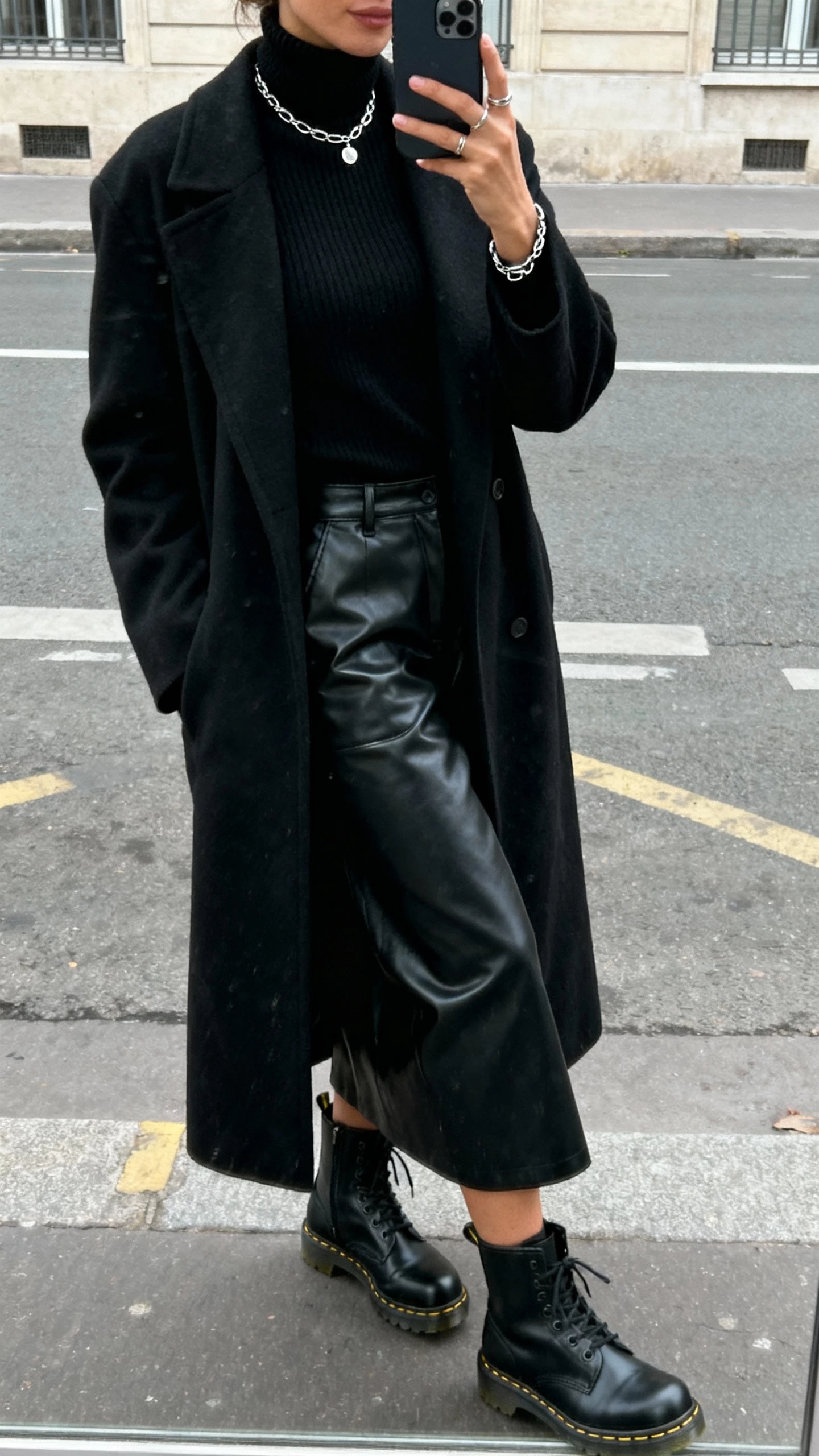 iPhone street reflection selfie of a woman in an all-black look mixing textures: black turtleneck, black coated or leather-look bottom, black long coat, black Doc Martens, subtle silver jewelry, face not visible, overcast daylight, casual iPhone photo.