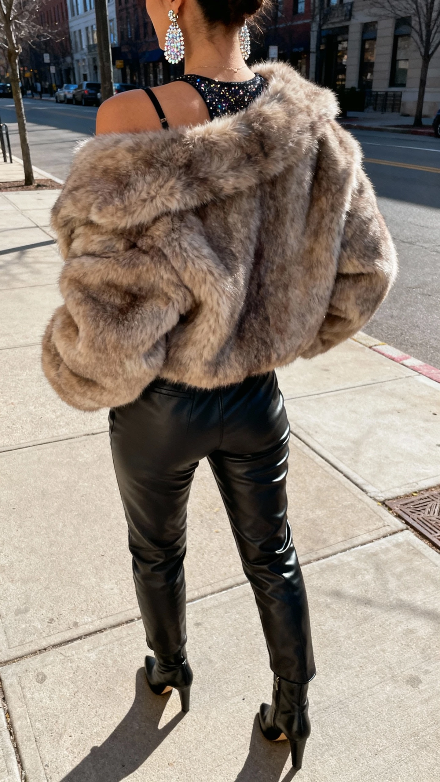 iPhone street-side back-view selfie of a woman in a faux-fur jacket, leather pants, and a party top with sparkly earrings and heeled boots, face not visible, city sidewalk in daylight, natural iPhone photo quality.