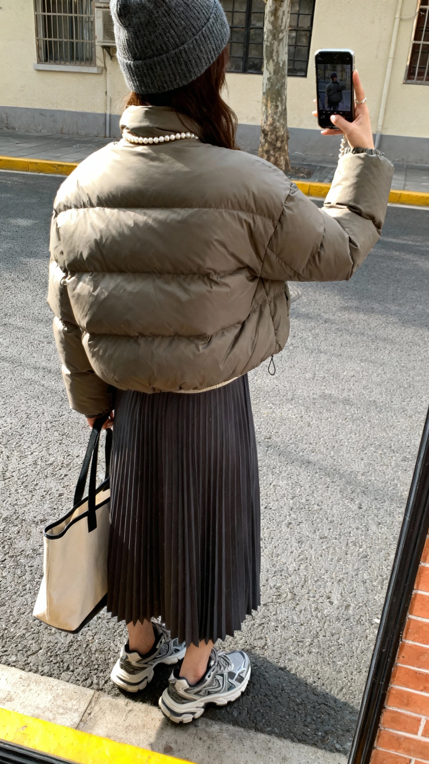 iPhone street-side back-view selfie of a woman wearing a puffer jacket, pearl necklace, pleated midi skirt, chunky sneakers, tote bag, beanie, face not visible, natural daylight, casual iPhone photo quality