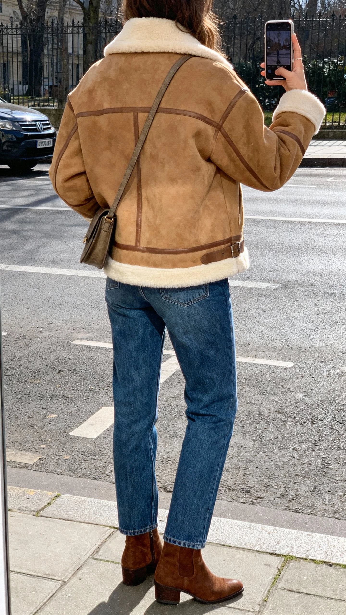 iPhone street-side back-view selfie of a woman wearing a tan shearling jacket with cream lining, straight blue jeans, brown ankle boots, crossbody bag, face not visible, winter daylight, casual iPhone photo.