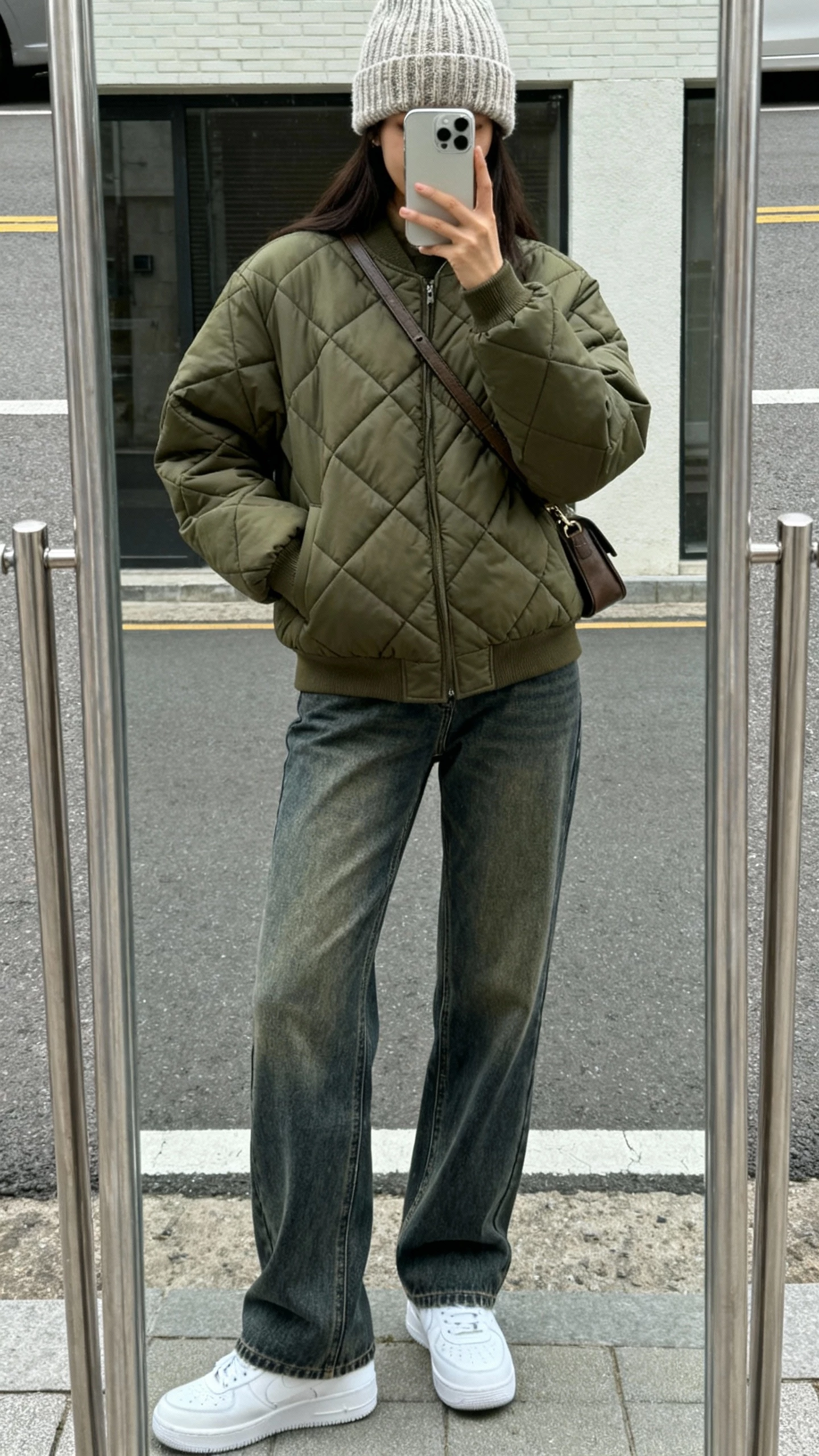 iPhone street-side mirror reflection selfie of a woman in an olive quilted jacket, straight-leg mid-wash jeans, clean white sneakers, knit beanie, crossbody bag, face not visible, overcast daylight, casual iPhone photo.
