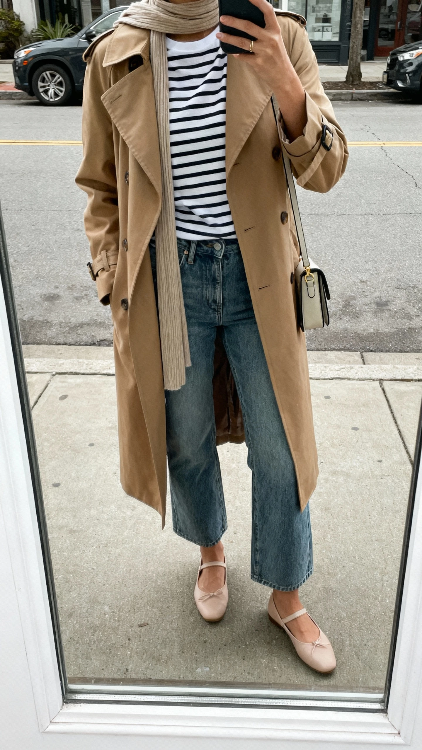 iPhone street-side mirror window selfie of a woman wearing a classic Breton striped tee, camel trench coat, medium-wash bootcut jeans, ballet flats, slim scarf and a small shoulder bag; face not visible, cropped at shoulders, overcast daylight, natural iPhone photo quality.