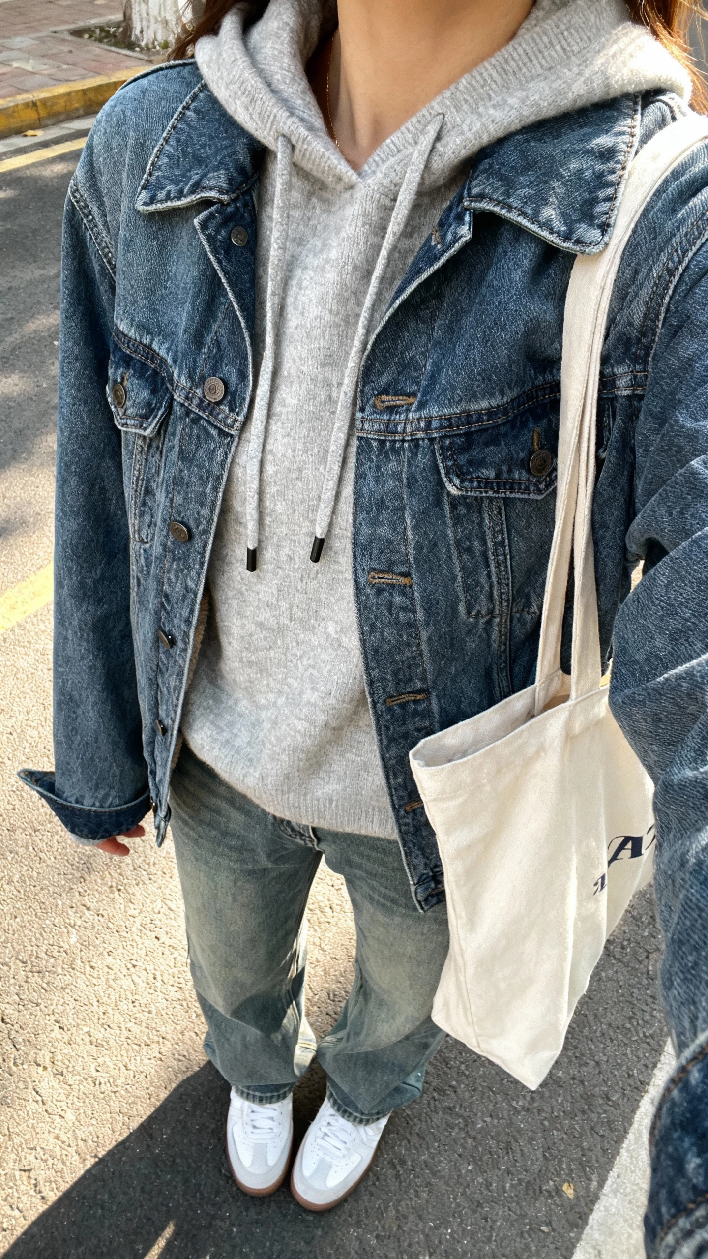 iPhone street-side selfie cropped at shoulders showing a woman wearing a medium-wash denim jacket layered over a light gray cashmere hoodie with drawstrings, relaxed jeans, white sneakers, canvas tote, face not visible, morning daylight, natural iPhone photo quality
