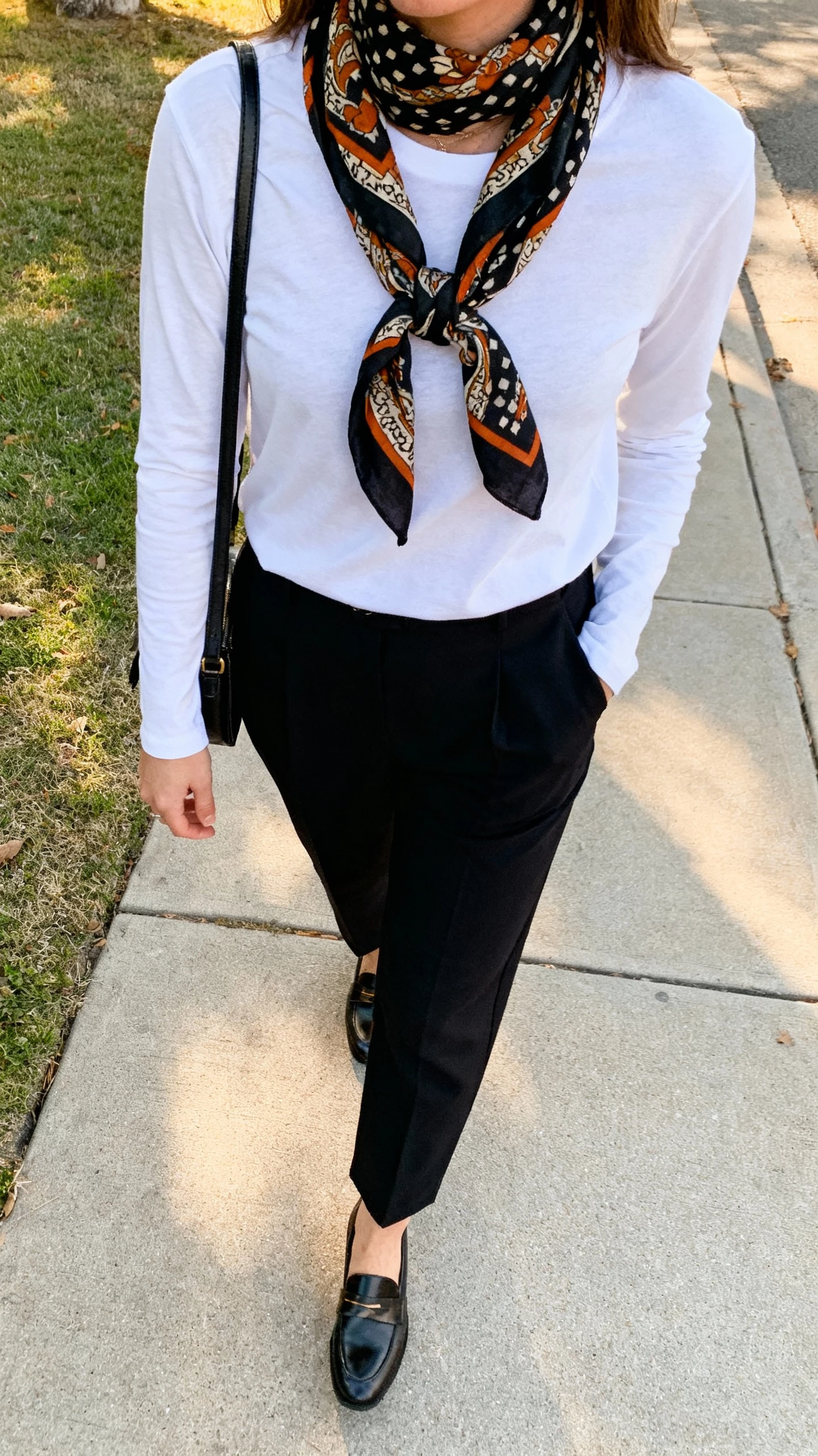 iPhone street-side selfie from chest down showing a woman in a minimal base (long-sleeve tee and tailored black trousers) with a bold statement scarf, loafers, crossbody bag, face not visible, sidewalk in soft afternoon light, natural iPhone photo quality.