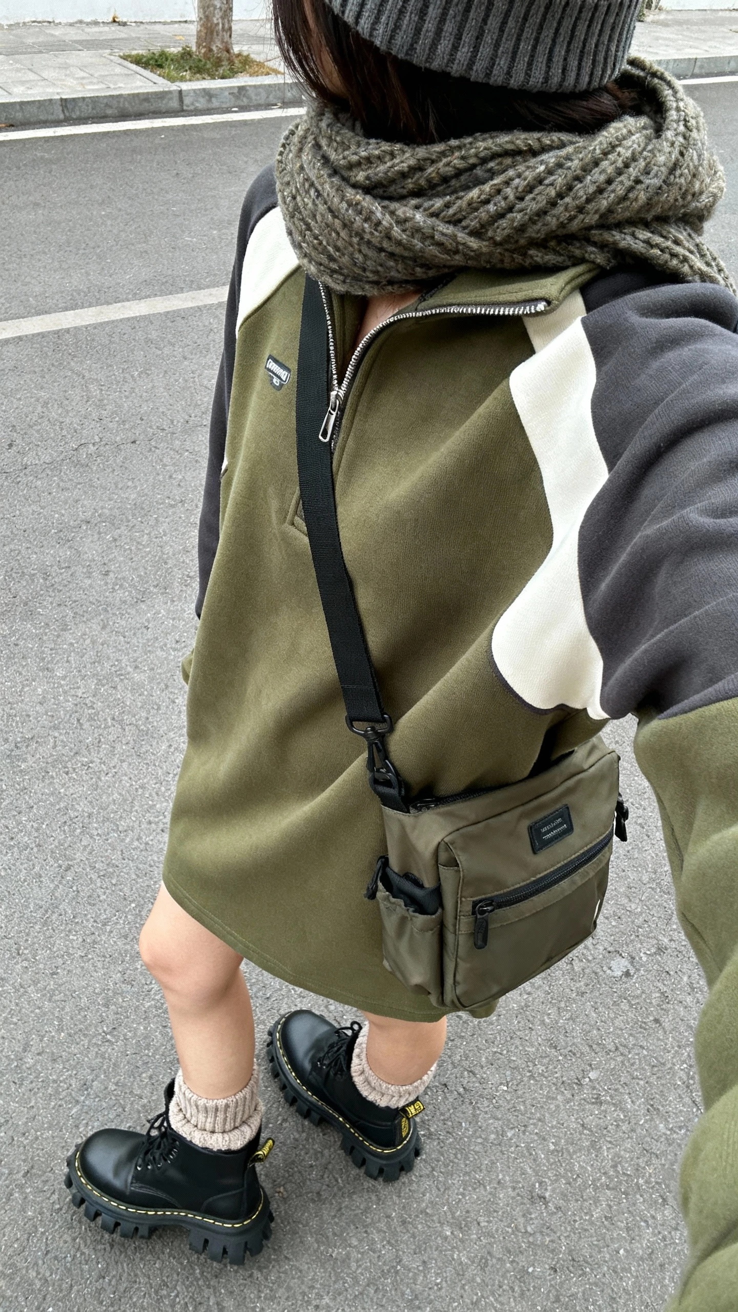 iPhone street-style back-view selfie of a woman wearing a relaxed half-zip sweater dress (olive/charcoal/cream) with lug-sole ankle boots or combat boots, sporty crossbody or utility messenger bag, ribbed beanie, chunky scarf, cozy wool socks peeking; face not visible, over-shoulder angle, overcast daylight, natural iPhone photo quality.