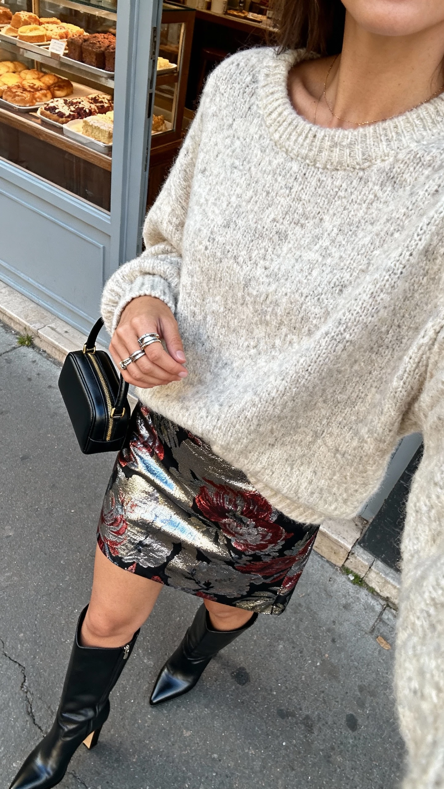 iPhone street-style selfie cropped at neck of a woman wearing a statement skirt (bold print or metallic) with a chunky knit sweater, heeled boots, layered rings, and a mini top-handle bag, face not visible, sidewalk outside a bakery with soft afternoon light, natural iPhone photo quality.