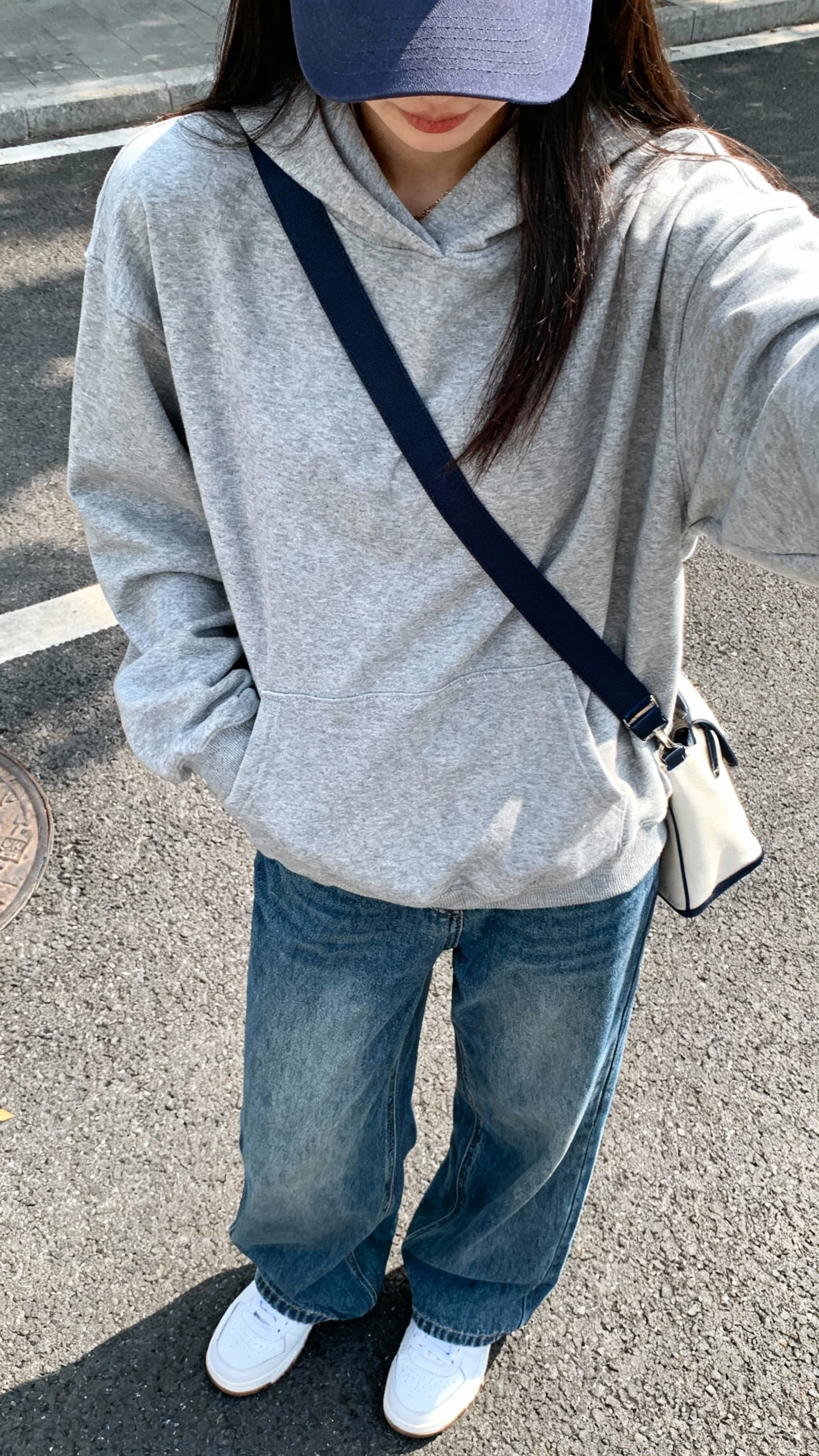 iPhone street-style selfie cropped at neck showing a woman in a gray hoodie with relaxed-fit blue jeans, white trainers, baseball cap, crossbody bag, face not visible, outdoor daylight, natural iPhone photo.